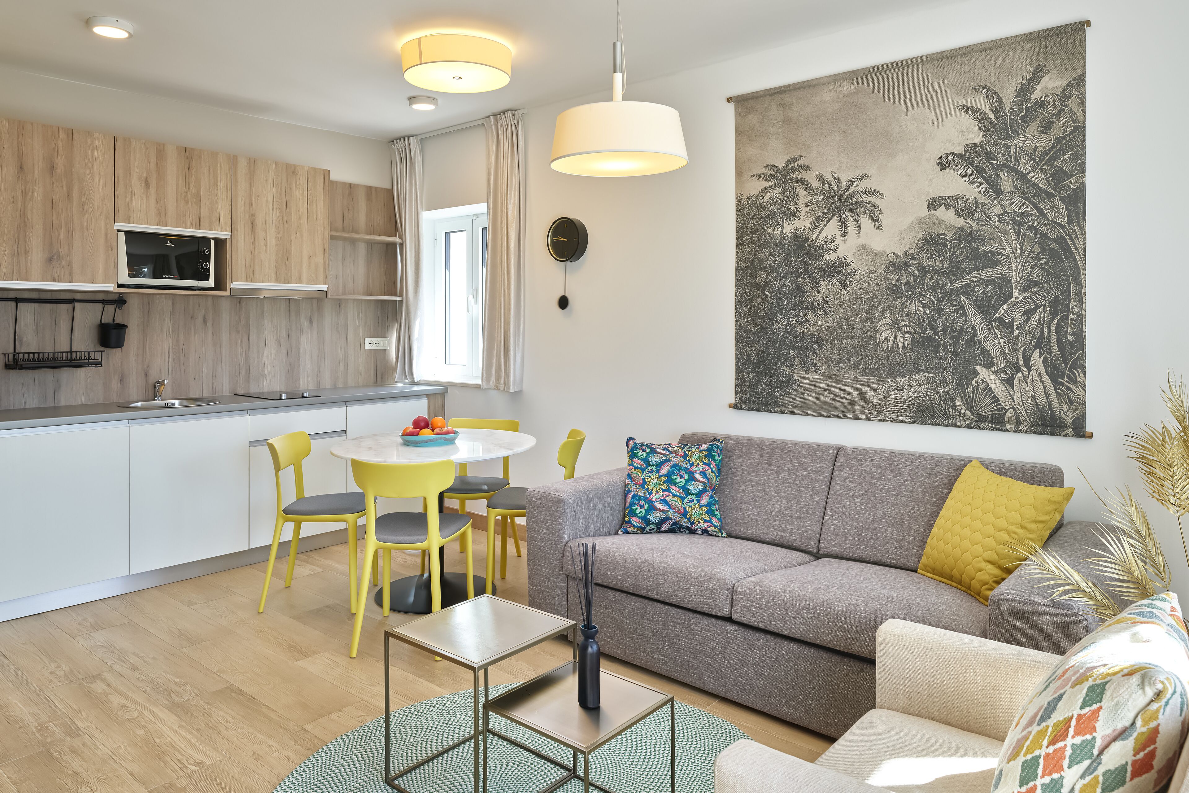 Een moderne woonkamer met een grijze bank, gele stoelen, een kleine tafel en een grote tropische muurschildering.