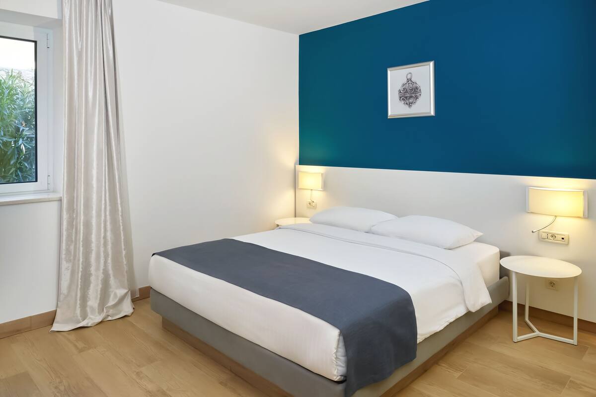 Een slaapkamer met een bed, nachtlampjes, een raam met gordijnen en een blauwe accentmuur.