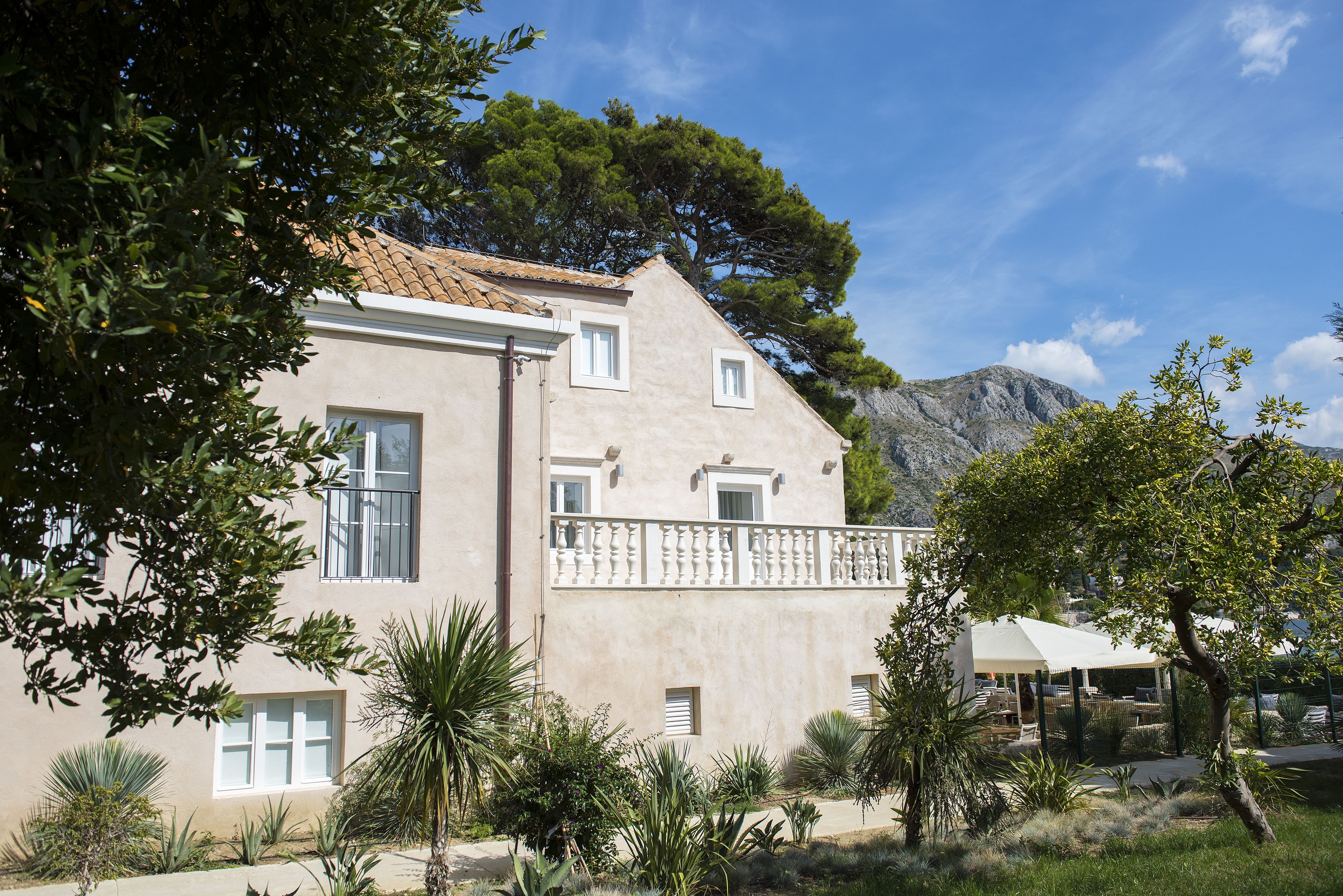 A two-story building with a balcony, surrounded by various plants and a mountainous backdrop.