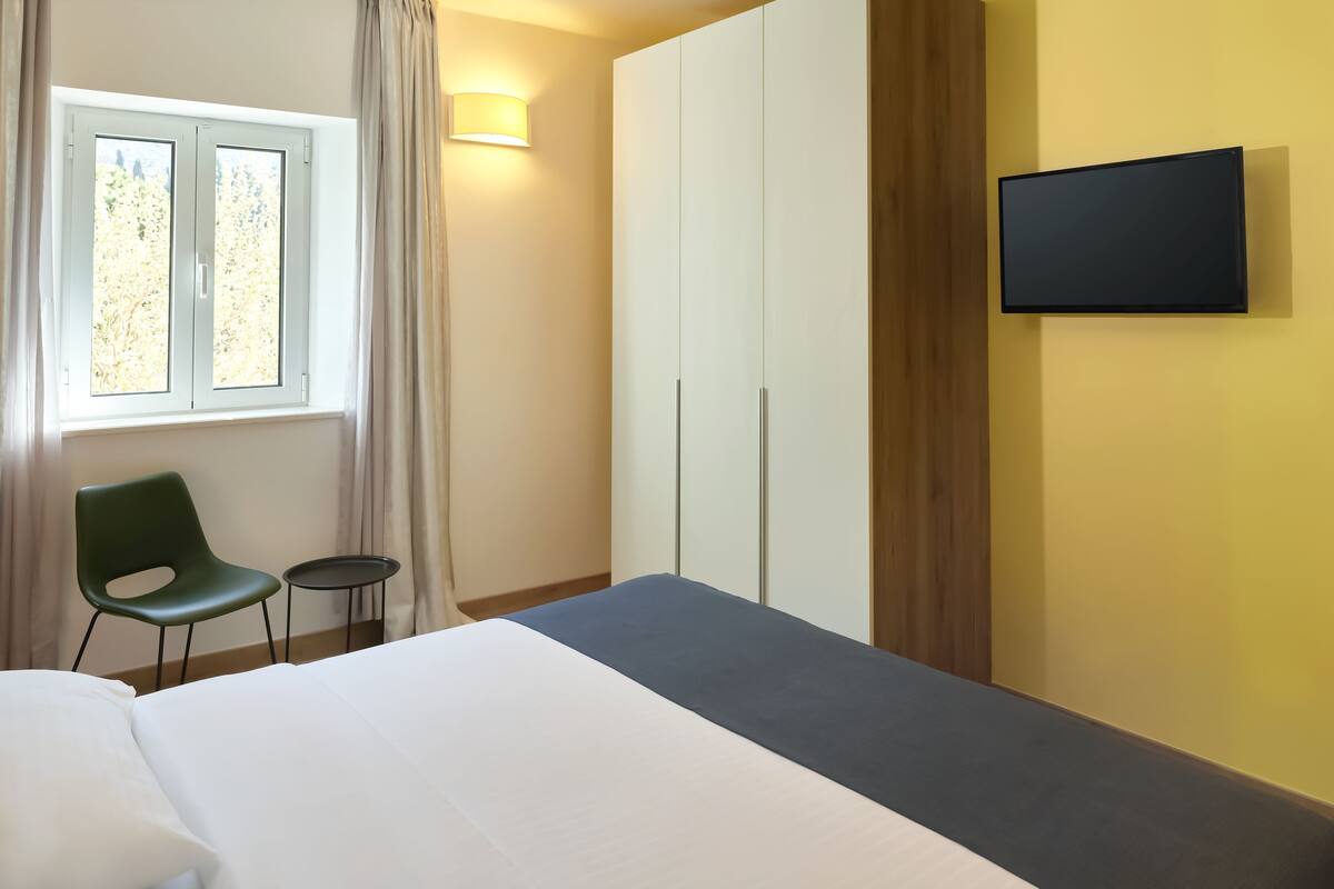 Een hotelkamer met een bed, een stoel, een klein tafeltje, een kledingkast en een aan de muur bevestigde televisie.