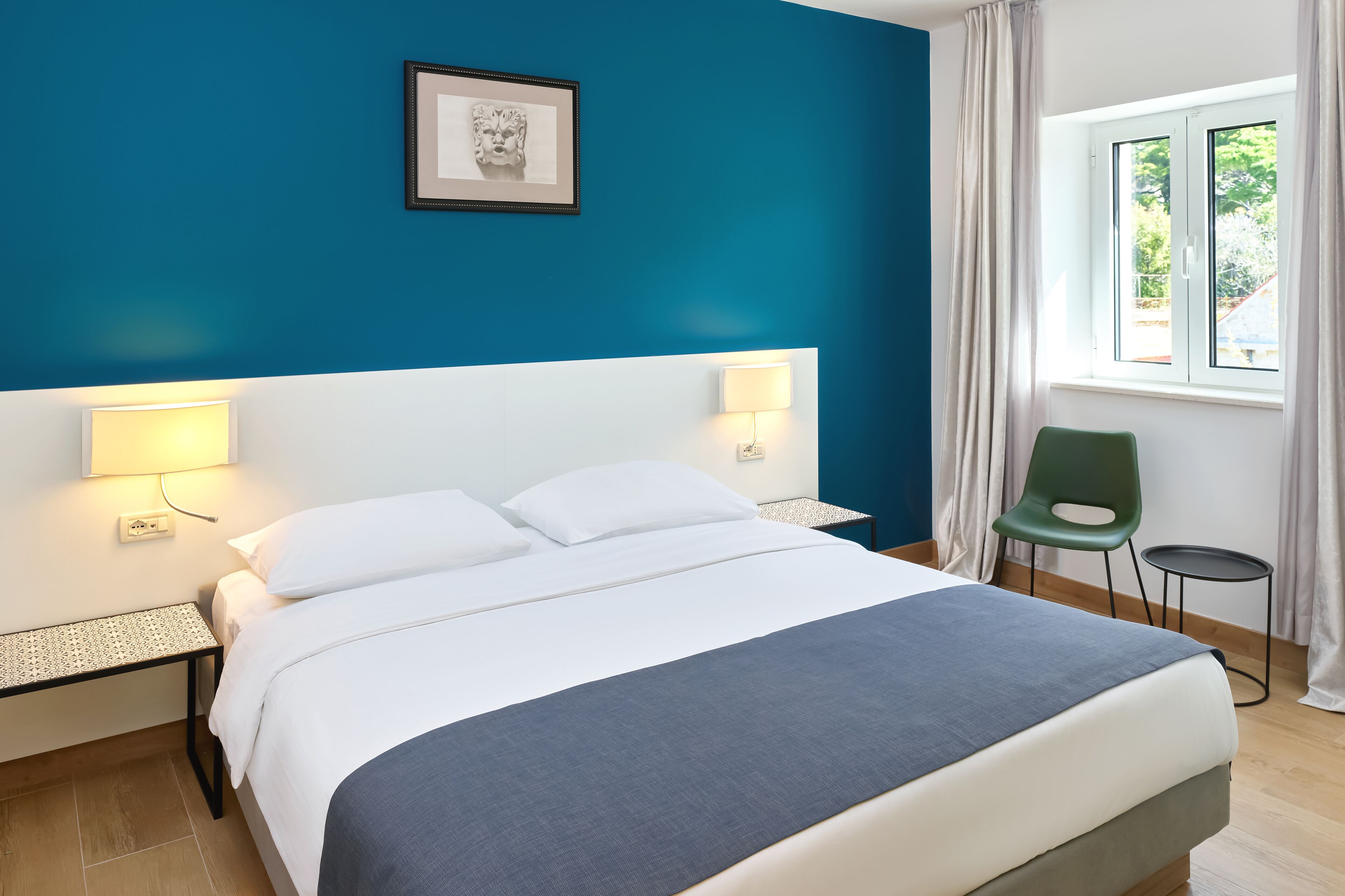 A hotel room with a bed, two bedside tables with lamps, a green chair, and a window with curtains.