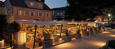 An outdoor dining area with tables and chairs, surrounded by potted plants and a building with a tiled roof.