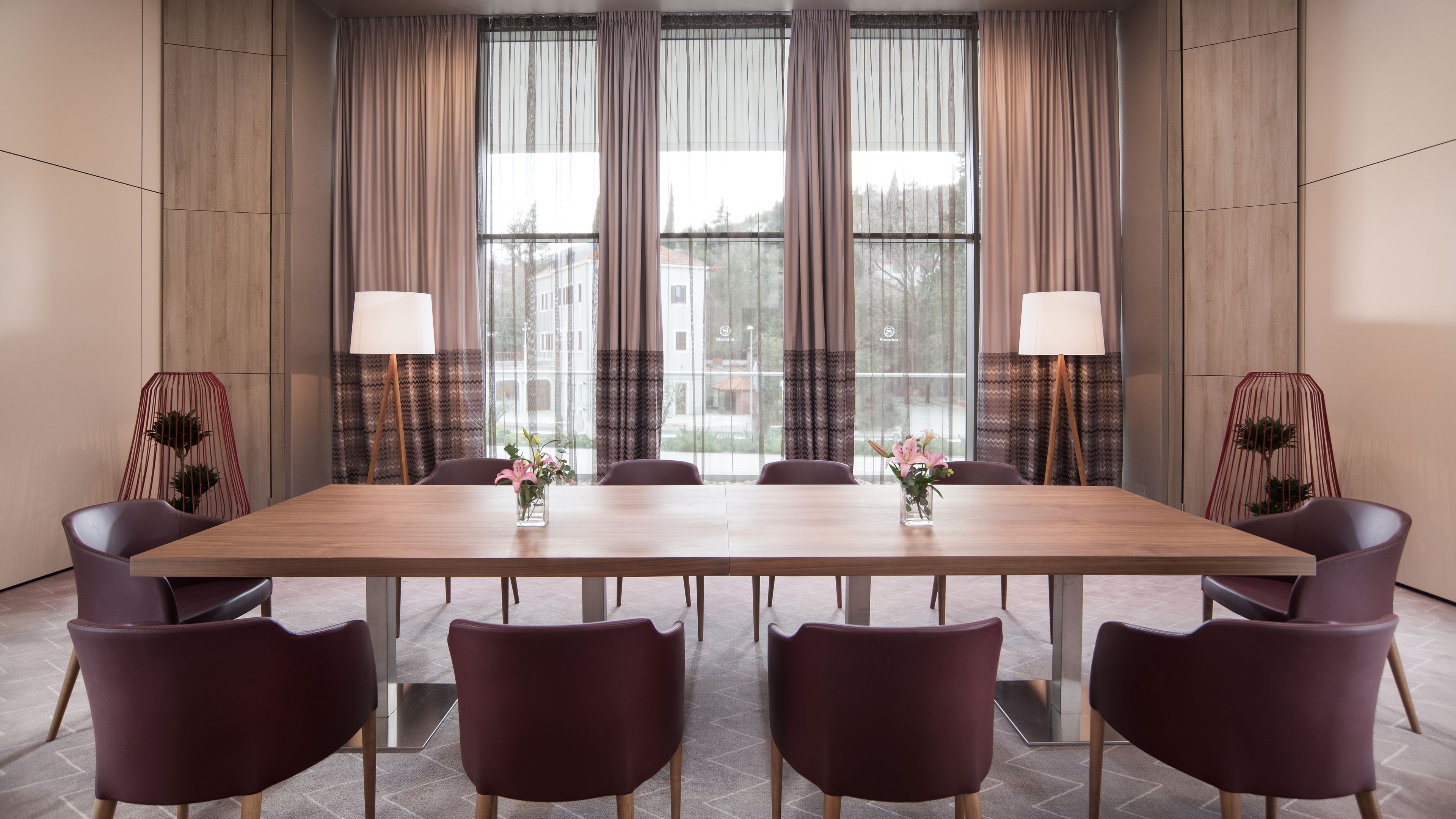 Un salón de conferencias con una mesa larga, seis sillas, dos lámparas de pie y vistas amplias a través de ventanas con cortinas.