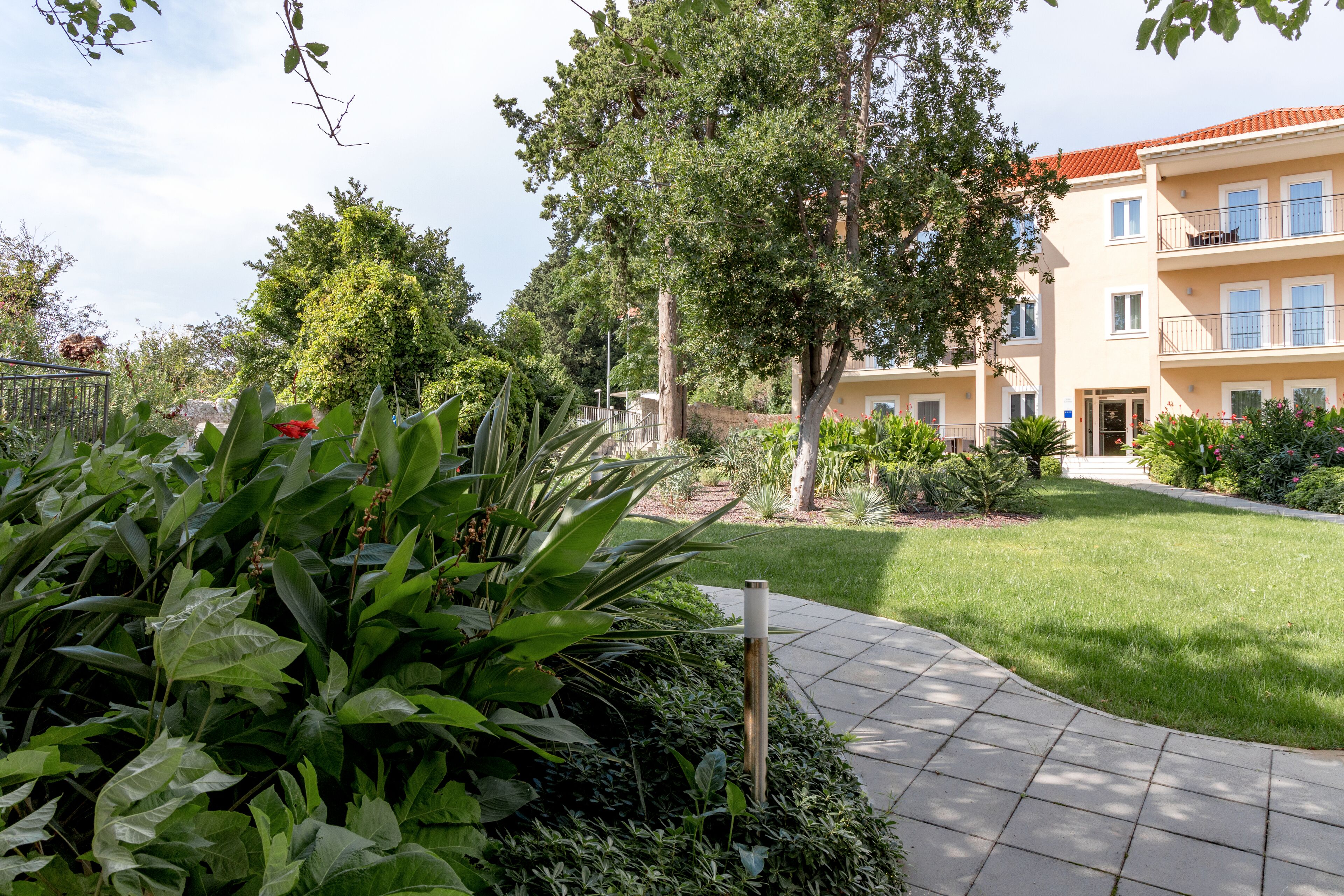 A courtyard with a paved walkway, lush greenery, and a multi-story building with balconies.