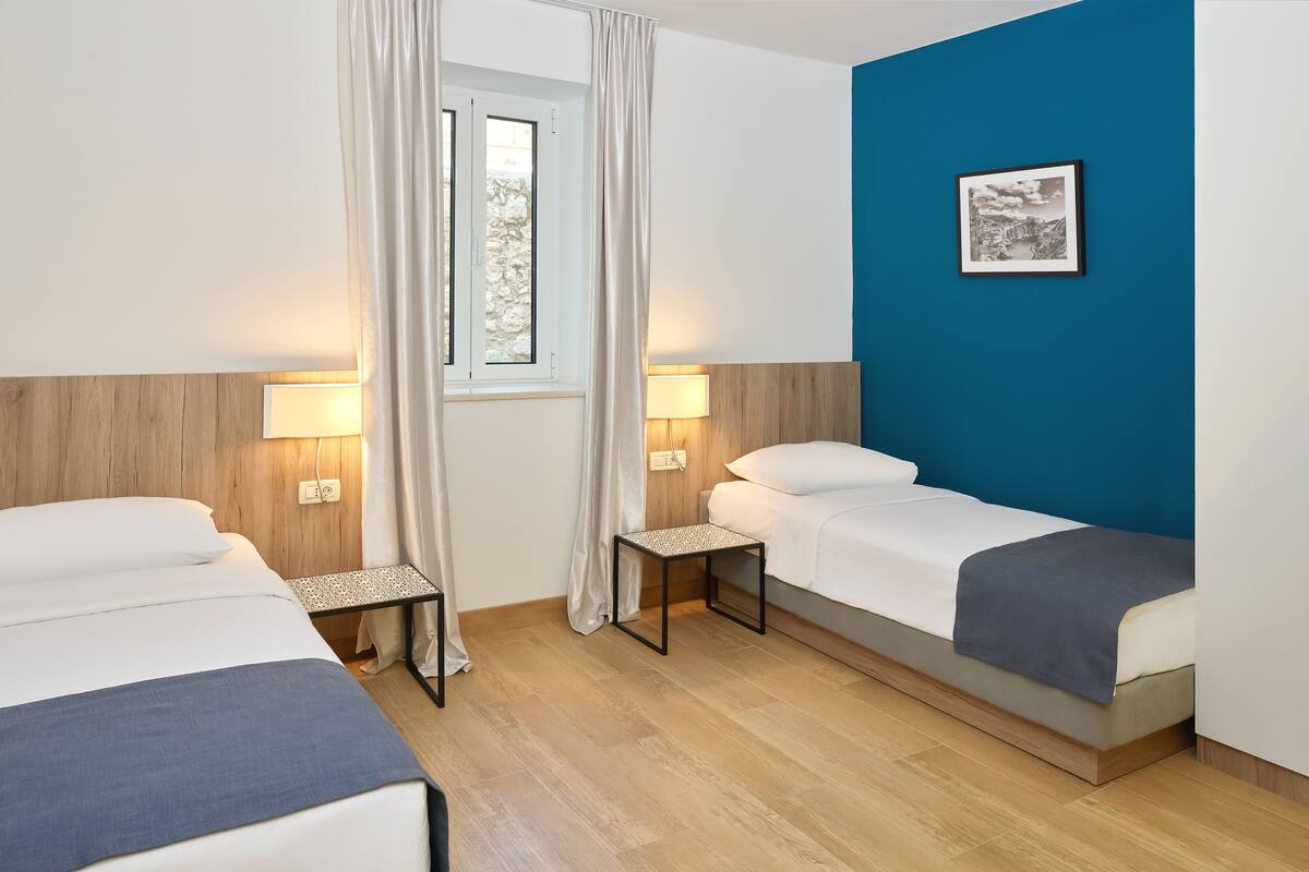 Een hotelkamer met twee enkele bedden, een klein tafeltje, een lamp, een raam met gordijnen en een ingelijst schilderij aan de muur.
