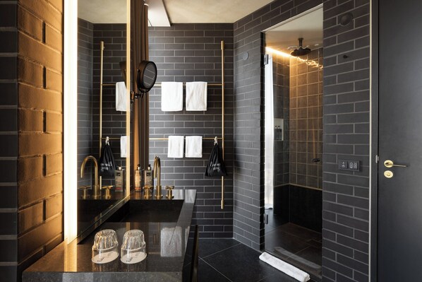 Un baño moderno con pared de ladrillo negro, un espejo grande, un lavamanos con detalles dorados y una zona de ducha.