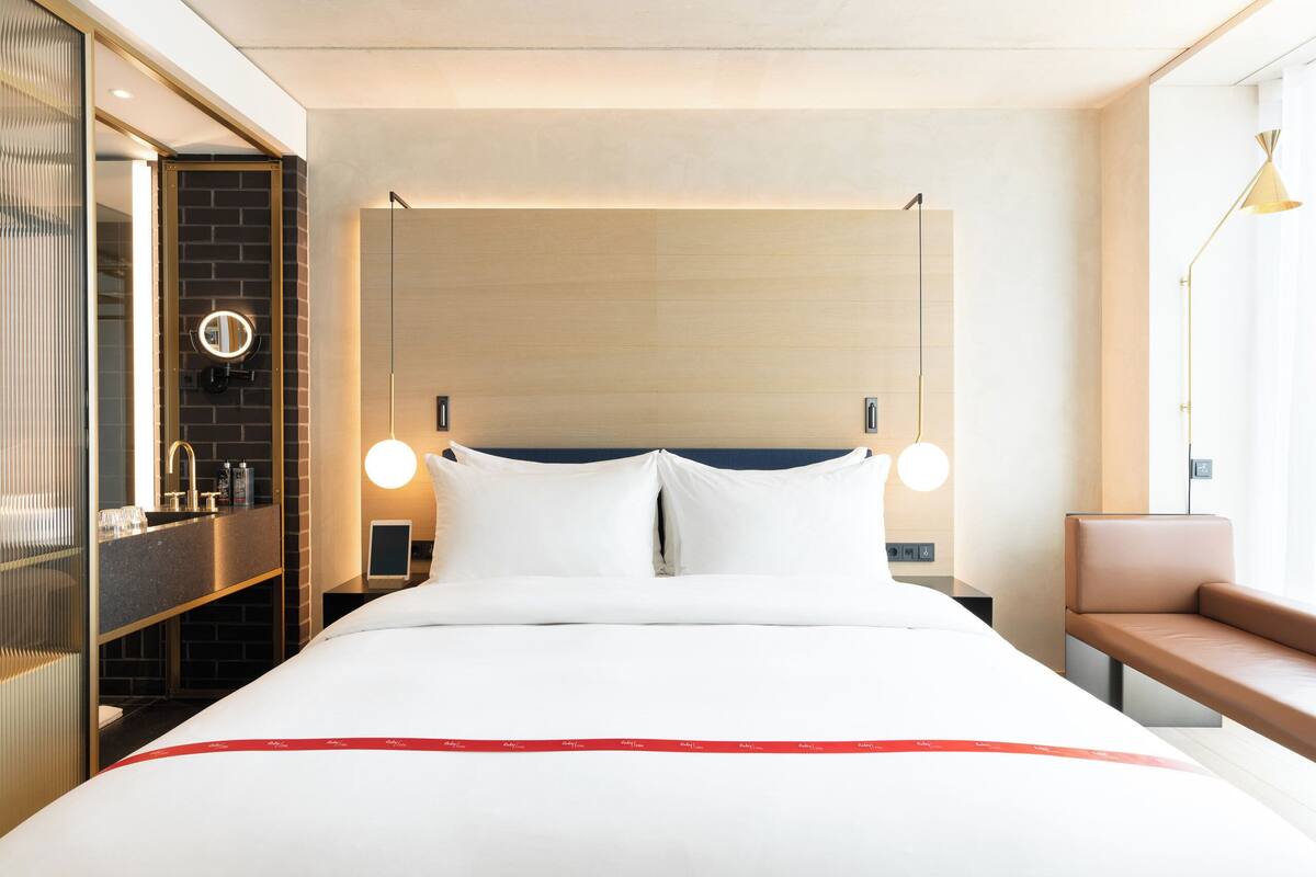 Une chambre d’hôtel avec un lit, une table de chevet, un miroir et un banc près de la fenêtre.
