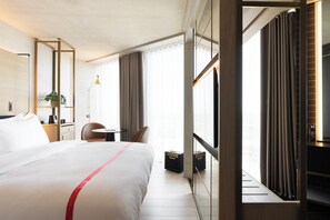 A hotel room with a large bed, a desk, a chair, a plant, and a window with curtains. - Ruby Emma Hotel Amsterdam By IHG (Amsterdam)