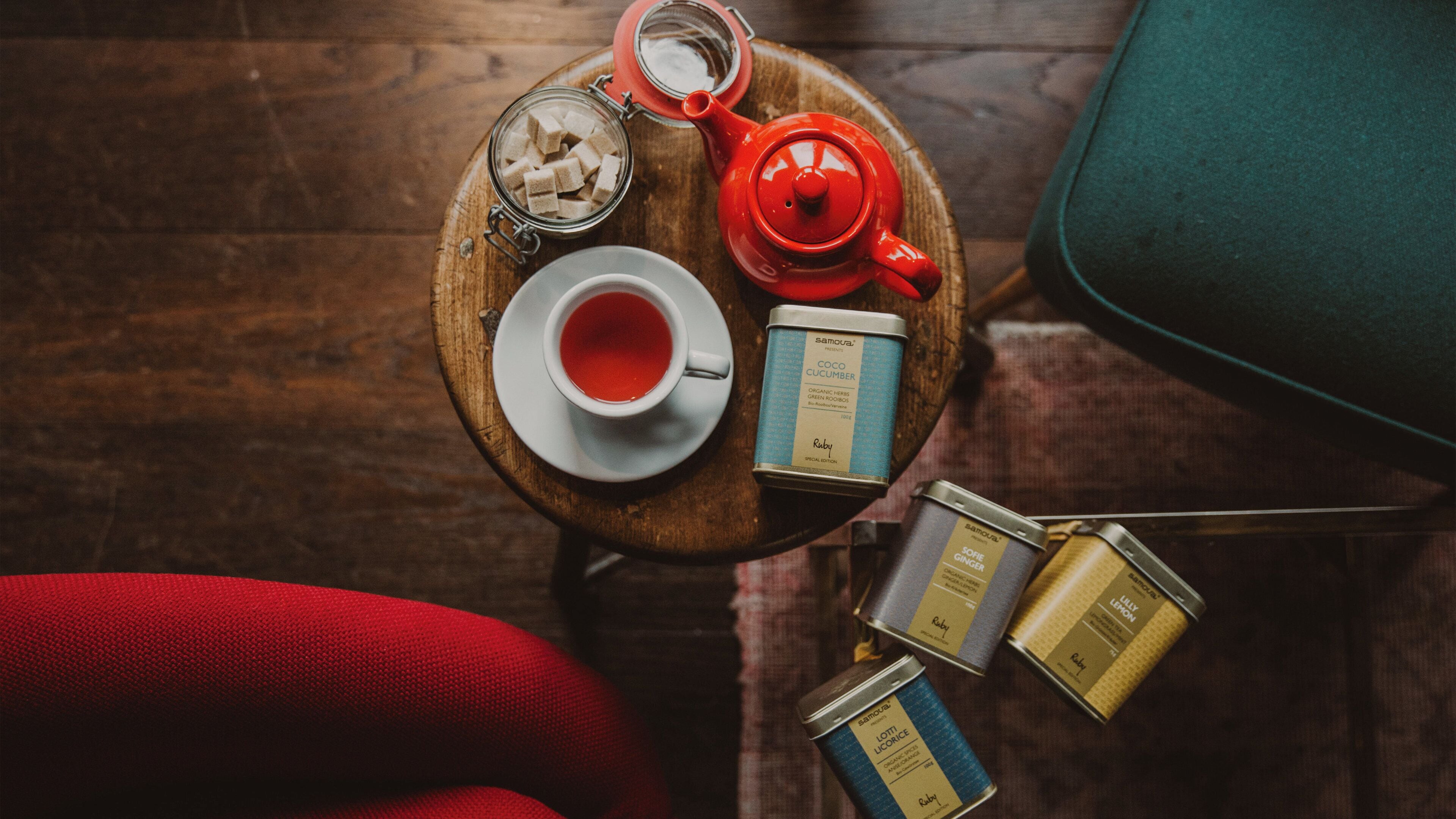A wooden table with a cup of tea, a red teapot, and several tins of Samouel products.
