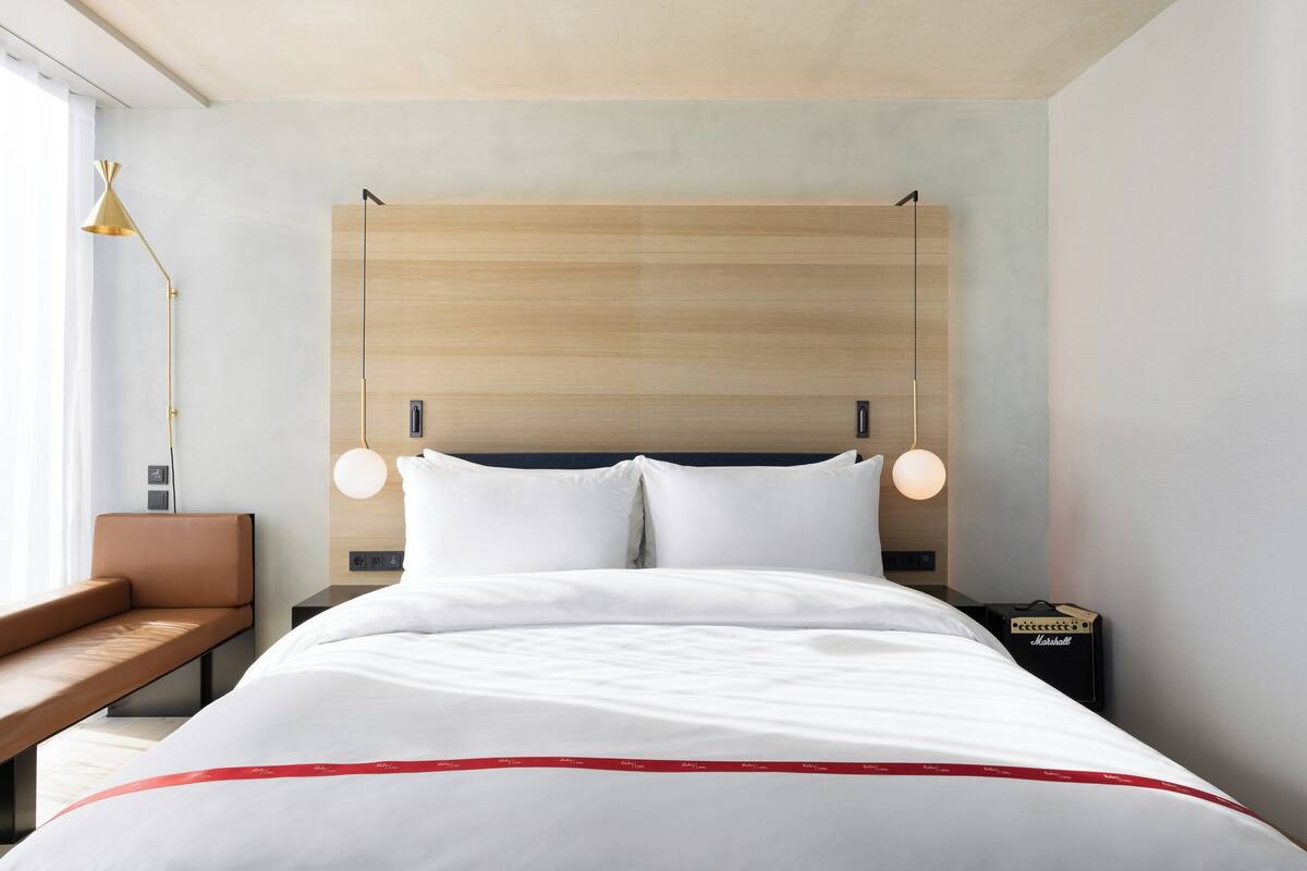 Une chambre d’hôtel moderne avec une tête de lit en bois, un lit avec une literie blanche, un banc, un bureau et une lampe.