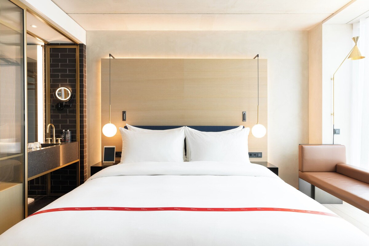 A hotel room with a bed, bedside table, mirror, and a bench by the window.