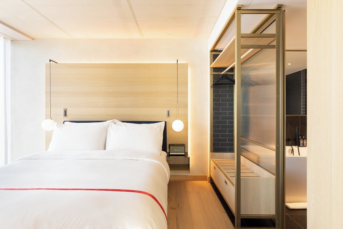 A modern hotel room with a large bed, a nightstand, a desk, and a glass partition.