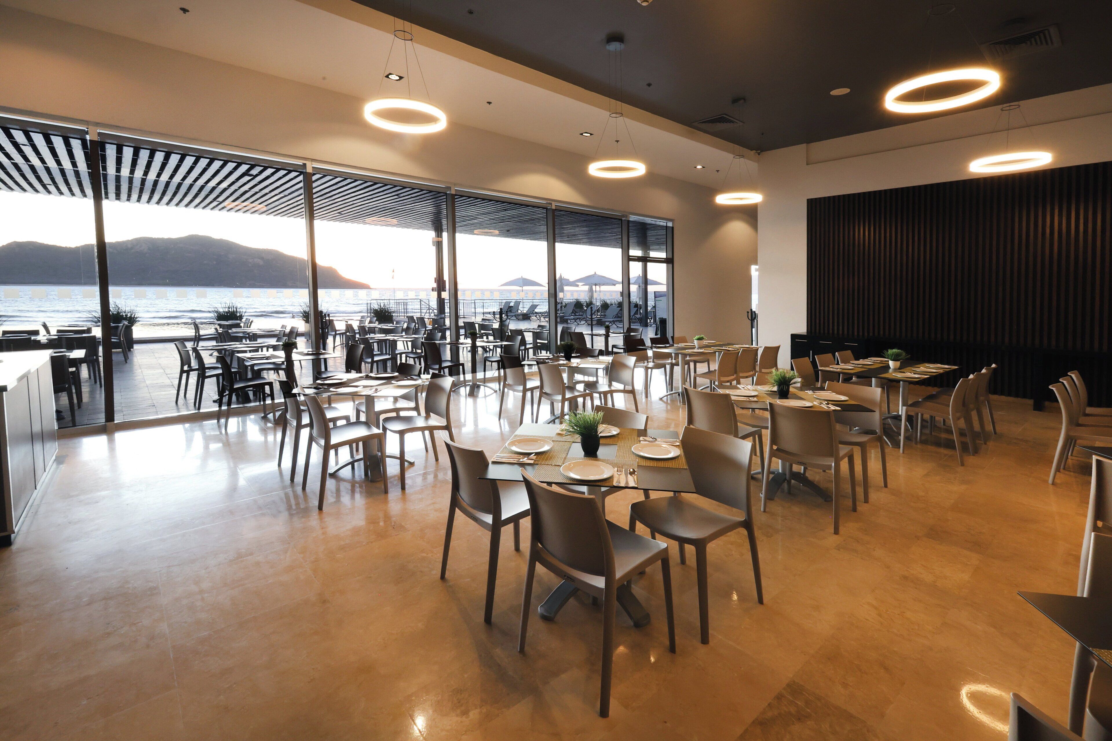 A spacious dining area with tables and chairs, large windows offering a view of the sea and mountains, and modern circular ceiling lights.