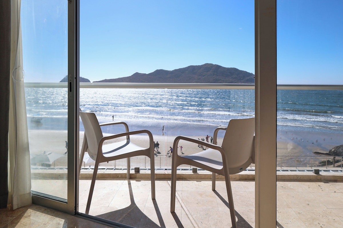 A room with a large glass door leading to a beach view. There are two white chairs and a small table in the room.