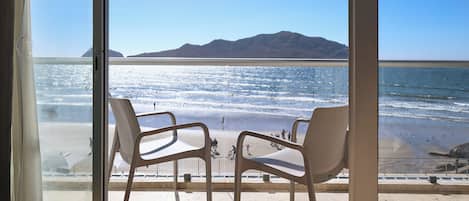 Una habitación con un gran ventanal que da a una vista de la playa. Hay dos sillas blancas y una mesita en la habitación.