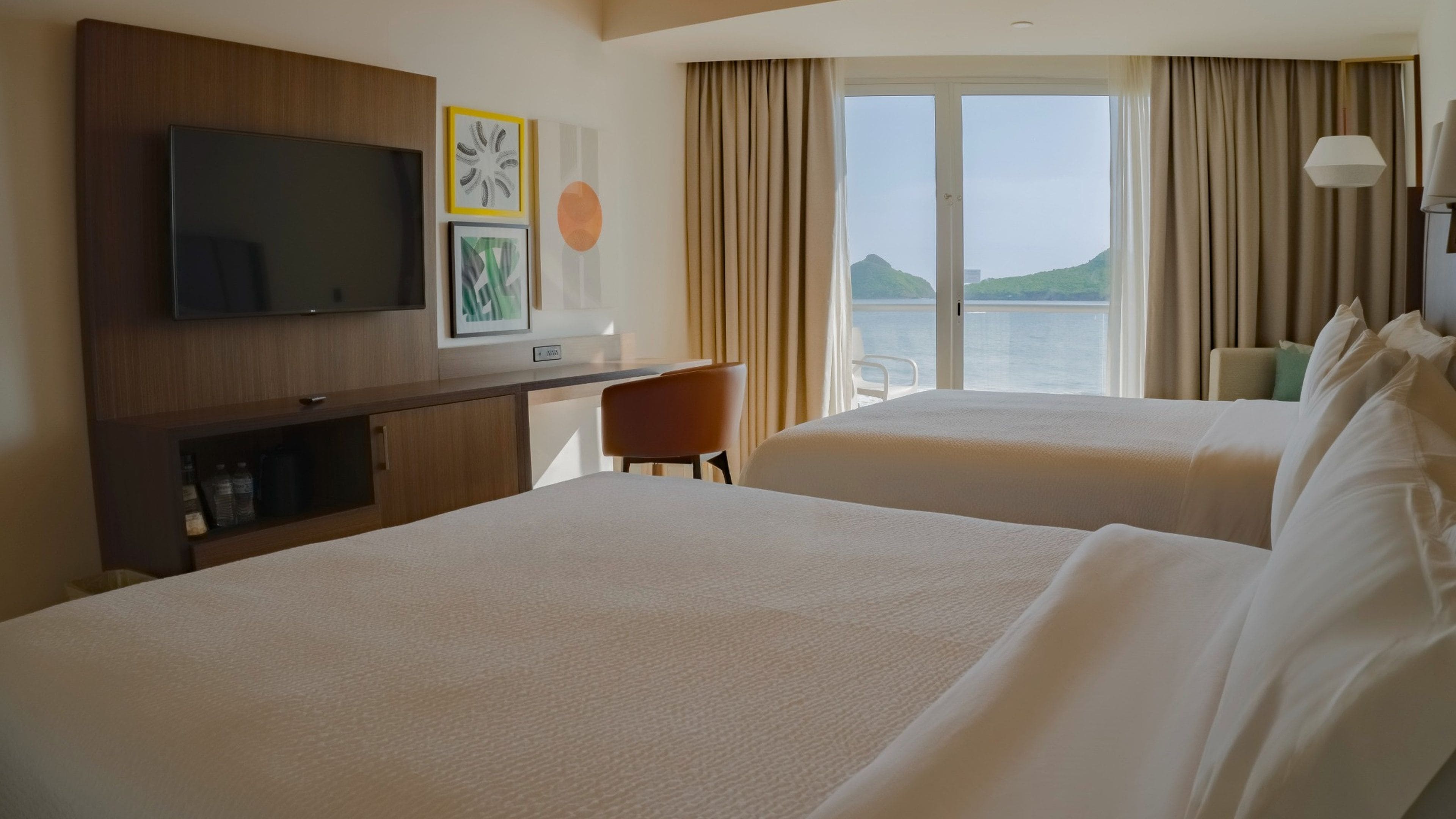 A hotel room with two beds, a desk, a television, and a window with curtains.