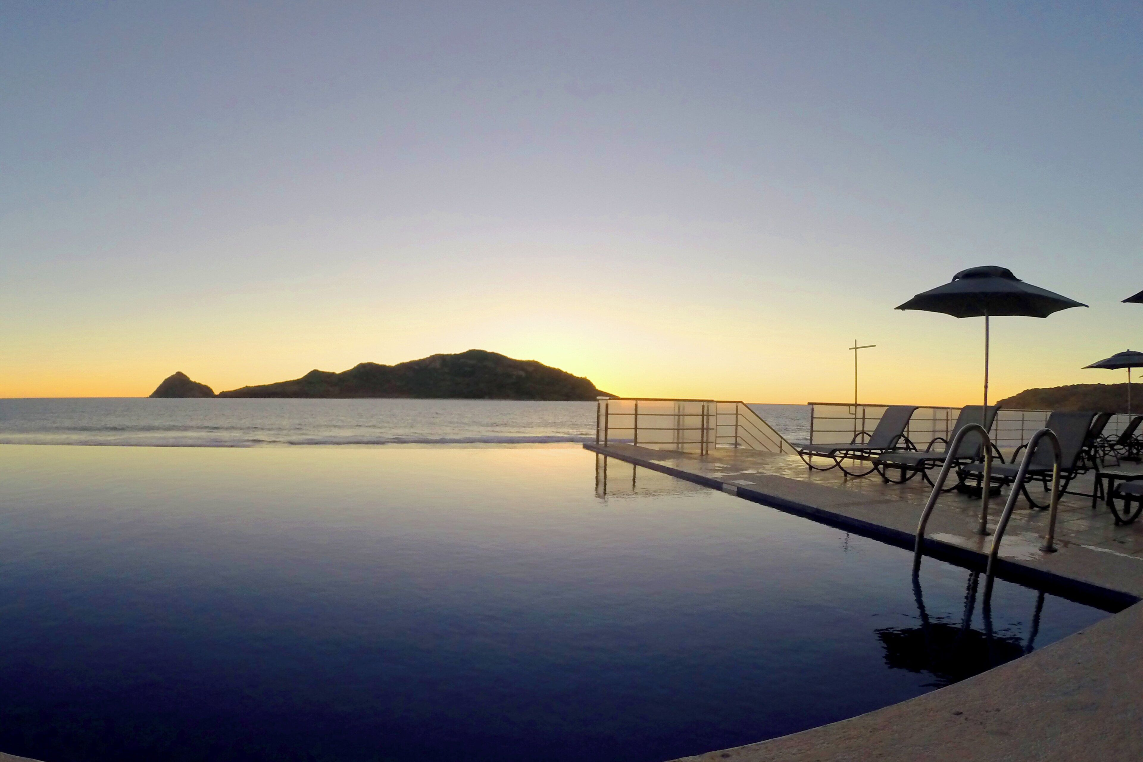 Une piscine à débordement offrant une vue sur l’océan et une île lointaine au coucher du soleil.