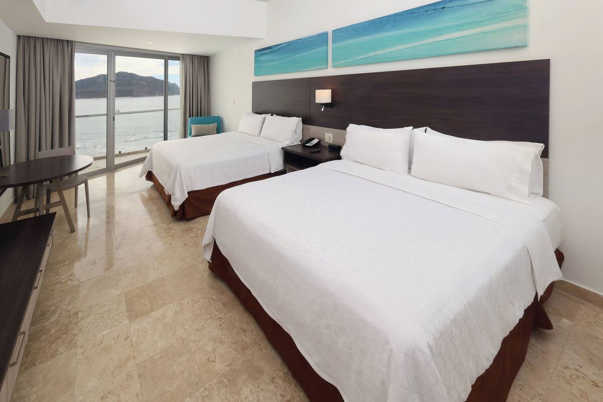 A hotel room with two beds, a desk, and a view of the sea.