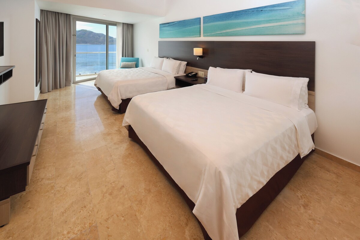 A hotel room with two beds, a desk, a TV, and a view of the sea.