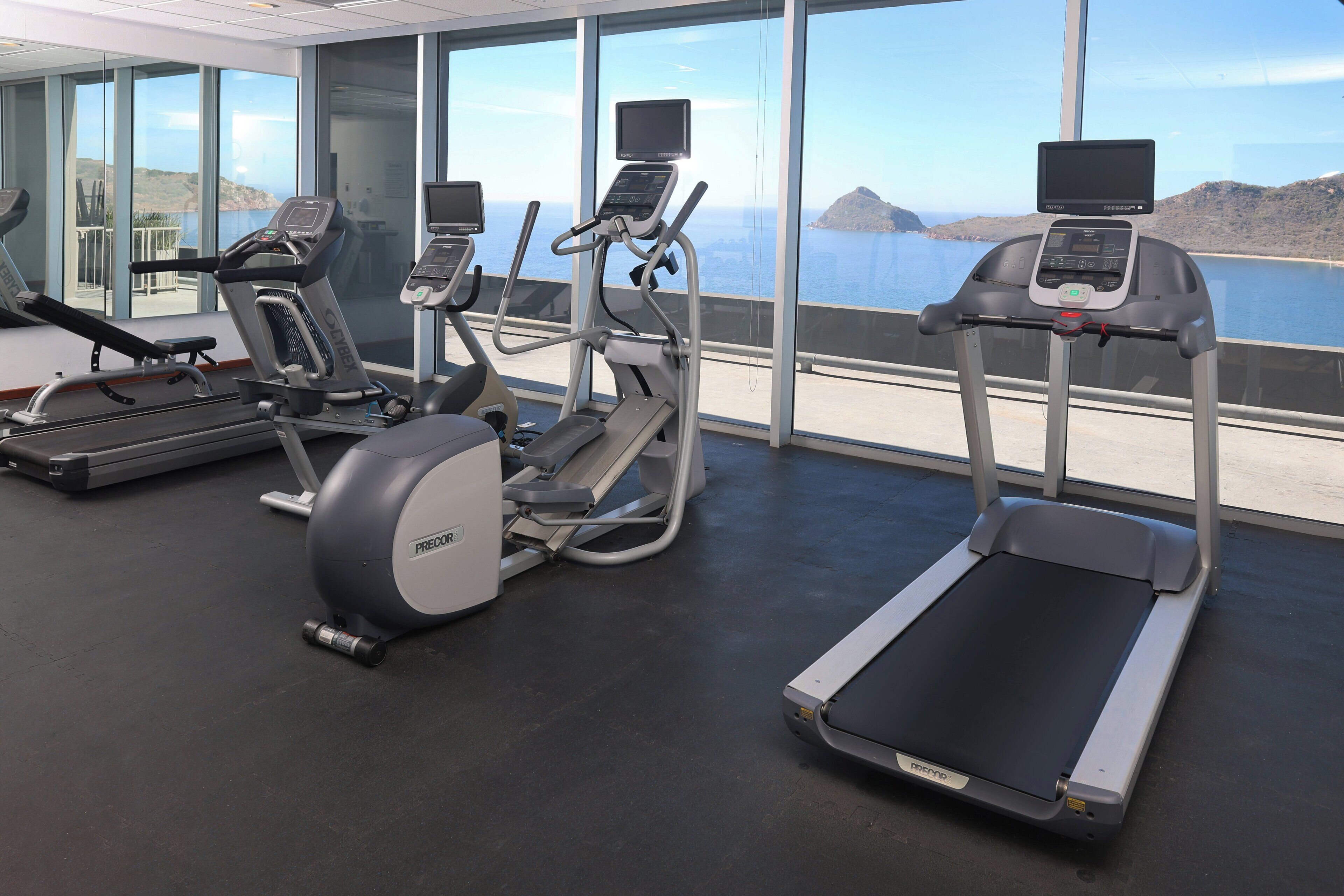 A gym with treadmills, an elliptical, and a stationary bike, overlooking a scenic view of the sea and hills.