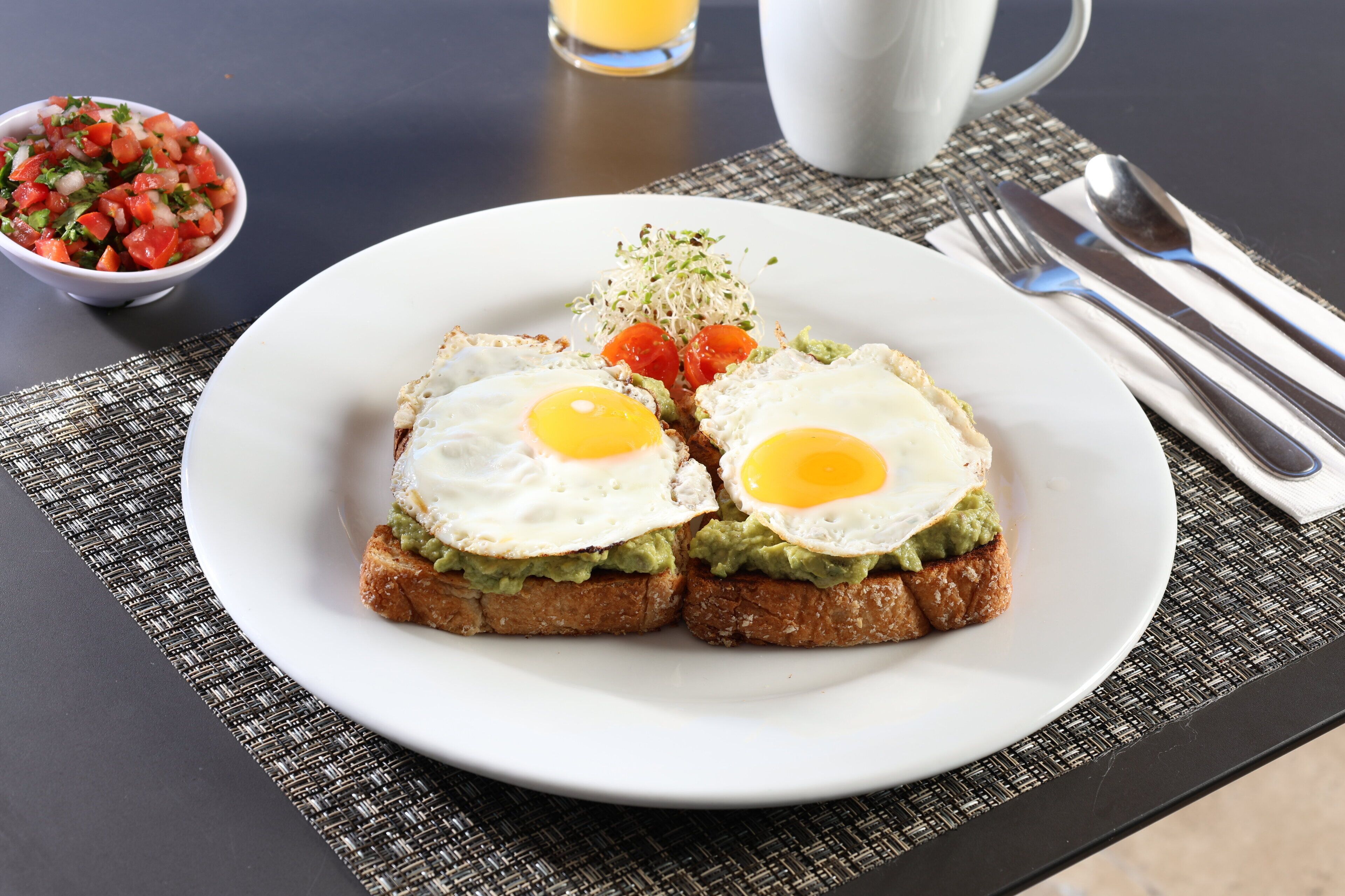 A breakfast plate with two fried eggs, avocado, and tomatoes on toast, accompanied by a bowl of salsa and a cup of coffee on a table.