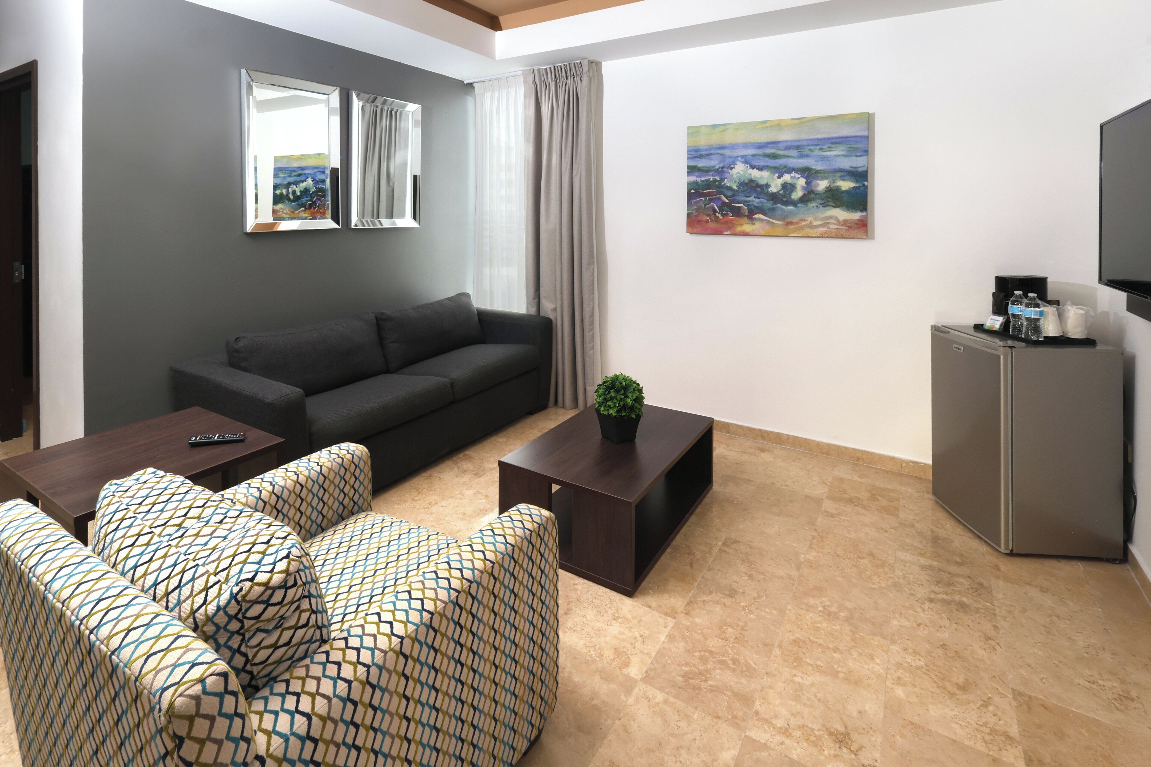 Un salon moderne avec un canapé gris foncé, des fauteuils à motifs, une table basse et une petite plante. On peut voir un téléviseur à écran plat et un réfrigérateur.