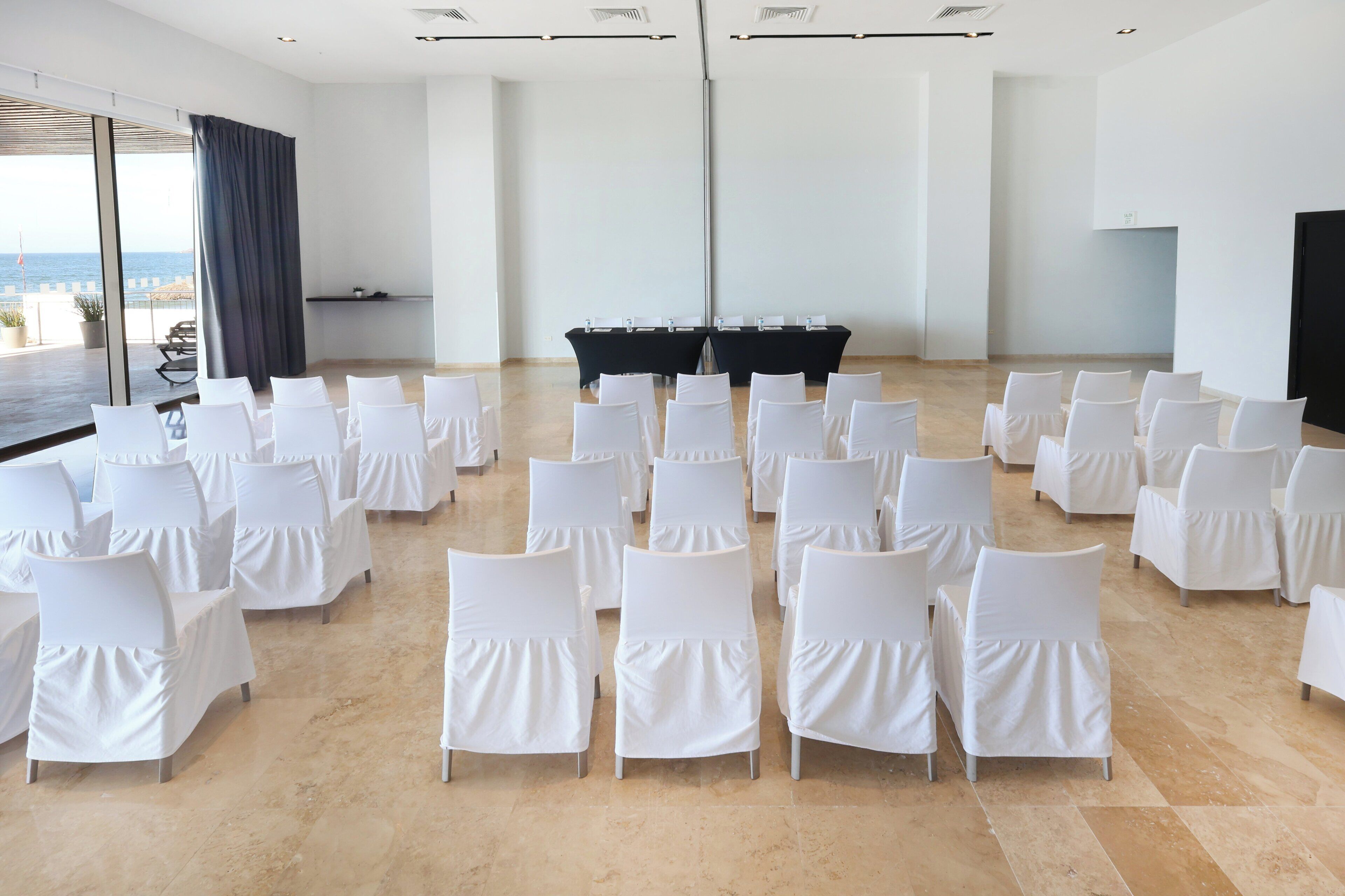 A conference room with white chairs arranged in rows, a black table in the center, and a large window with a view of the sea.