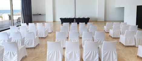 A conference room with white chairs arranged in rows, a black table in the center, and a large window with a view of the sea.