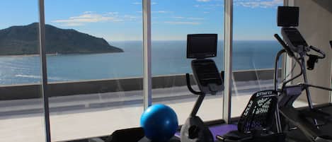 A gym with exercise equipment and a large window overlooking a body of water and a distant island.