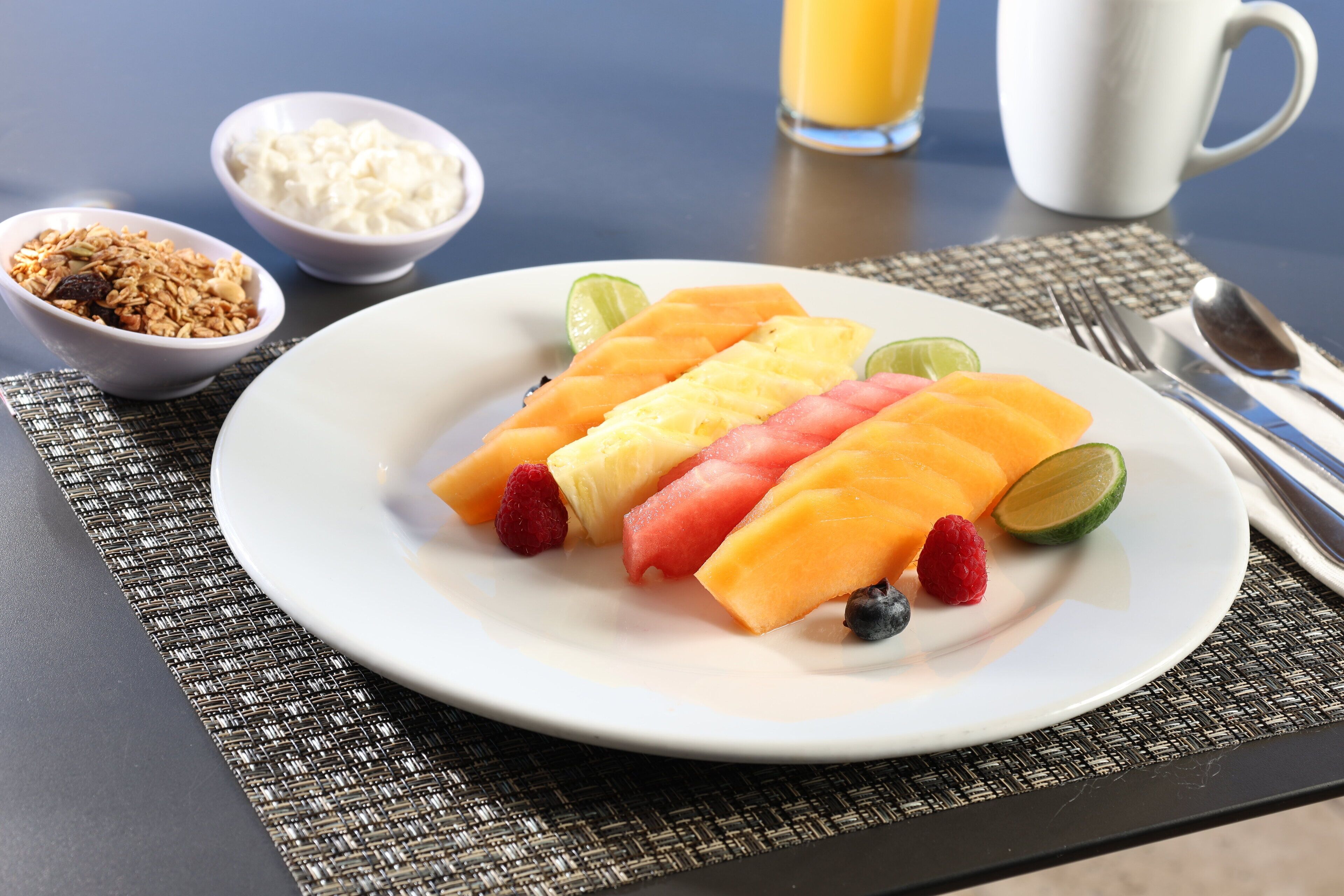 Une assiette de salade de fruits, un verre de jus d’orange et des bols de yaourt et de granola sont posés sur une table.