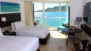 Habitació d'hotel amb dos llits, un balcó amb vista al mar, una taula petita amb una cadira i una televisió.
