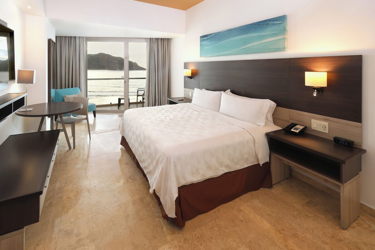 A hotel room with a bed, bedside table, desk, chair, and a view of the sea.
