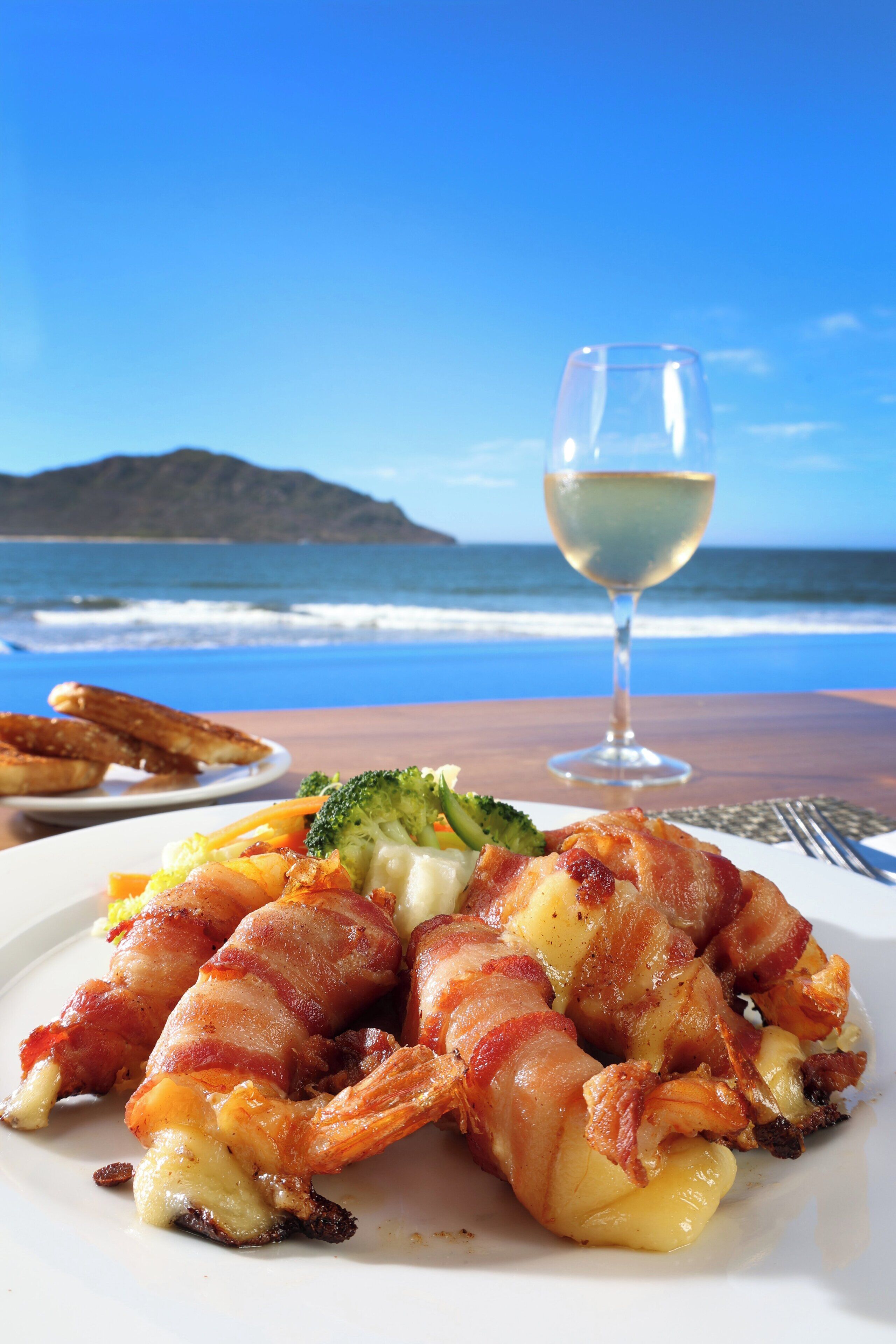 A plate of food with bacon-wrapped shrimp and broccoli, a glass of white wine, and a view of the ocean.