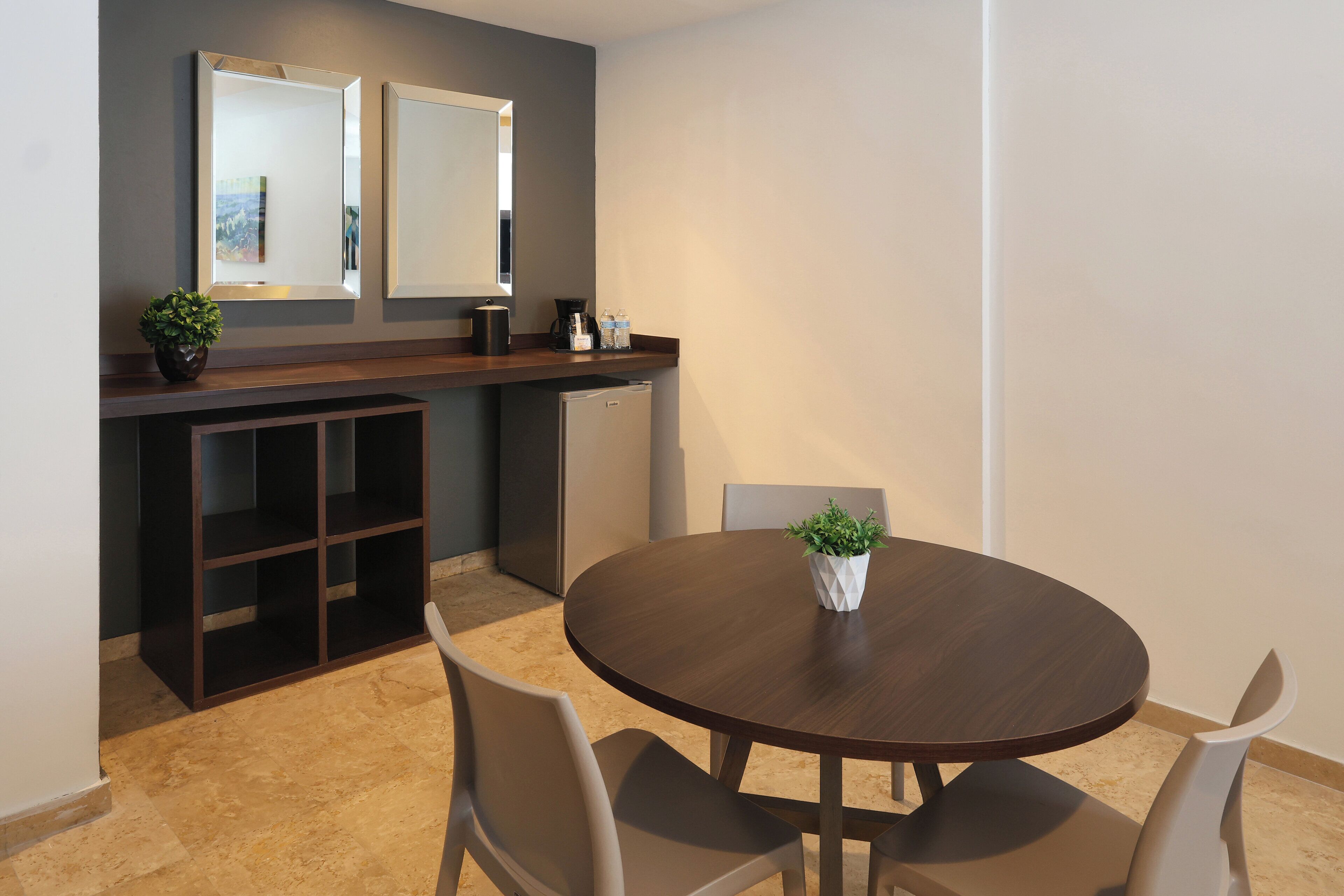 A dining area with a round table, two chairs, a potted plant, a coffee machine, and a refrigerator.