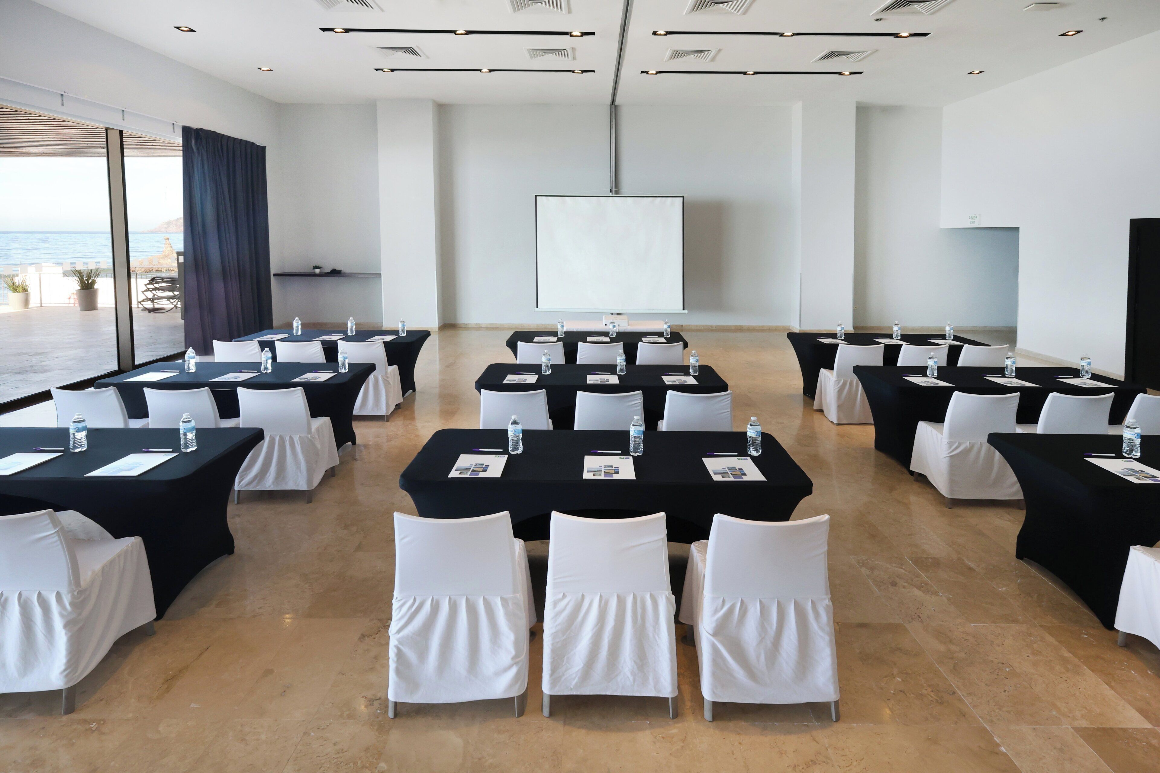 A conference room with multiple tables, white chairs, and a large screen.