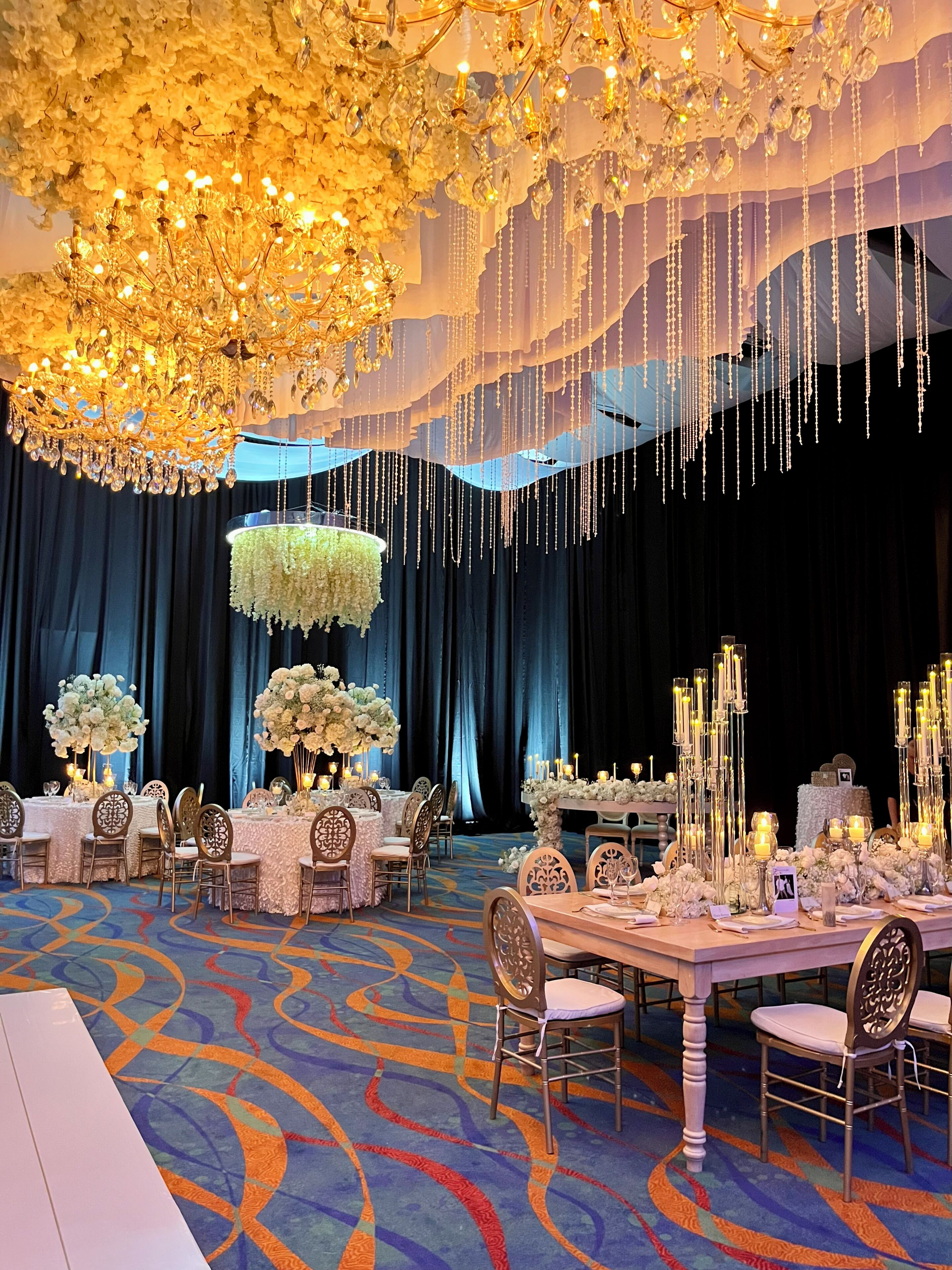 A banquet hall with tables set for a formal event, featuring chandeliers, floral arrangements, and draped curtains.