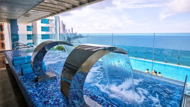 A rooftop infinity pool with a waterfall feature, overlooking a city skyline and the ocean.