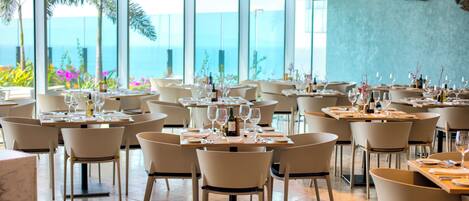 A restaurant with multiple tables and chairs, large windows with a view of the ocean, and decorative fish wall art.