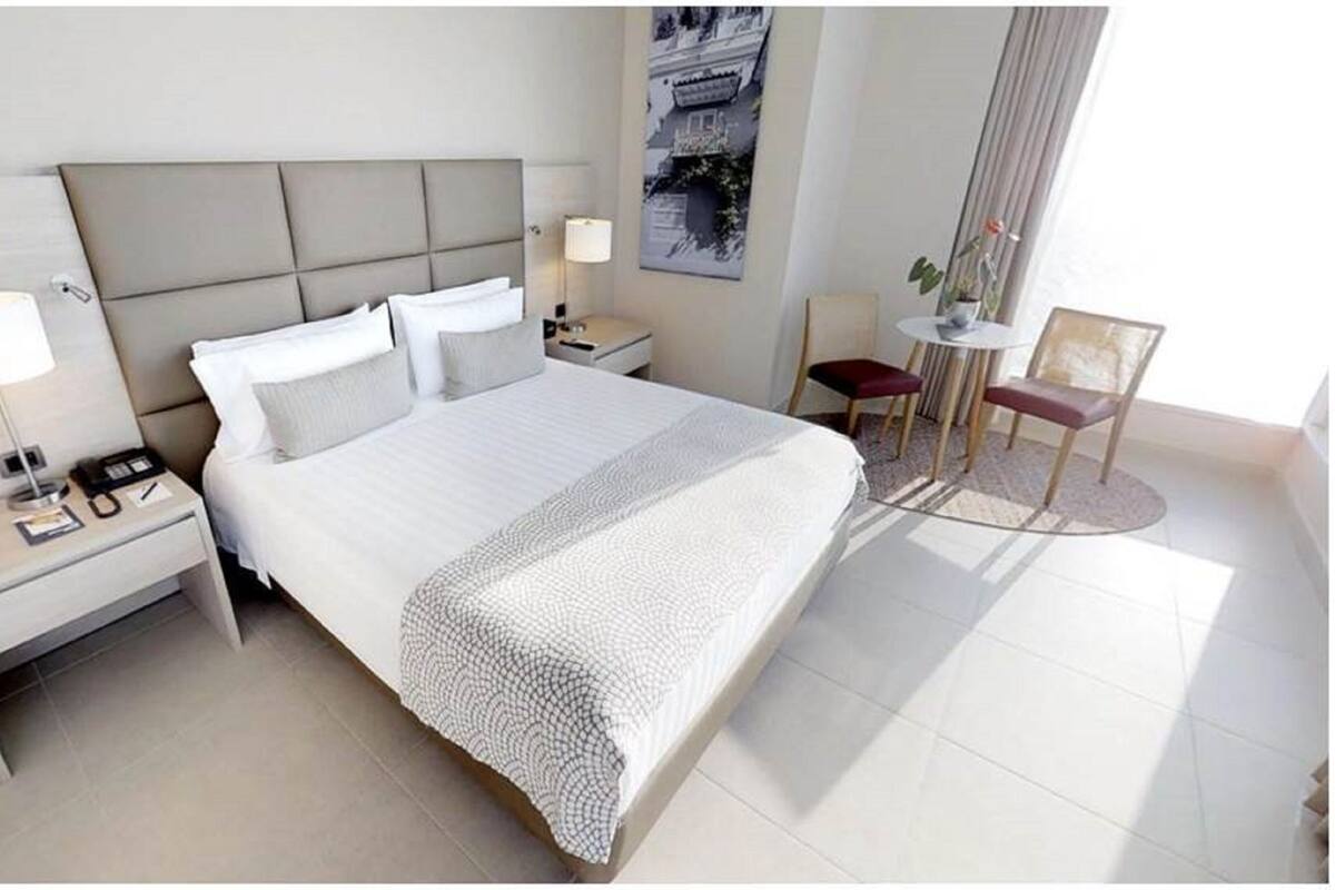 A hotel room with a large bed, a bedside table with a lamp, a small table with chairs, and a wall-mounted TV.