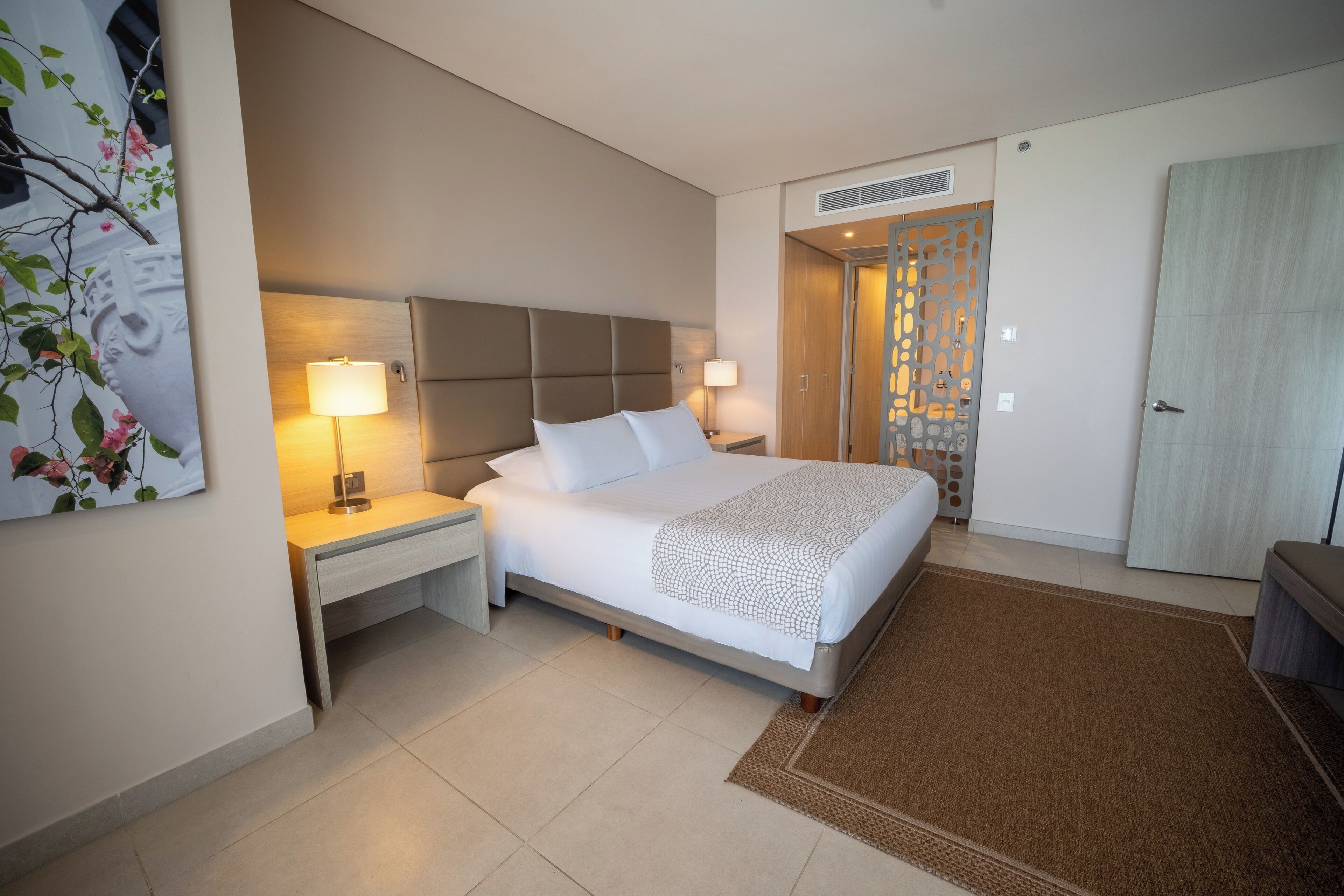 A hotel room with a bed, nightstands, a lamp, a door, and a wall decoration.