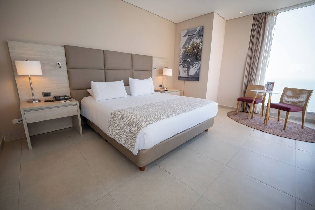 A hotel room with a large bed, bedside tables, a desk, a chair, and a window with curtains.
