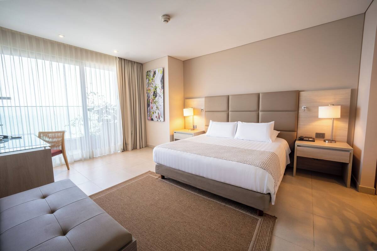 A hotel room with a large bed, two bedside tables with lamps, a desk, a chair, and a large window with curtains.