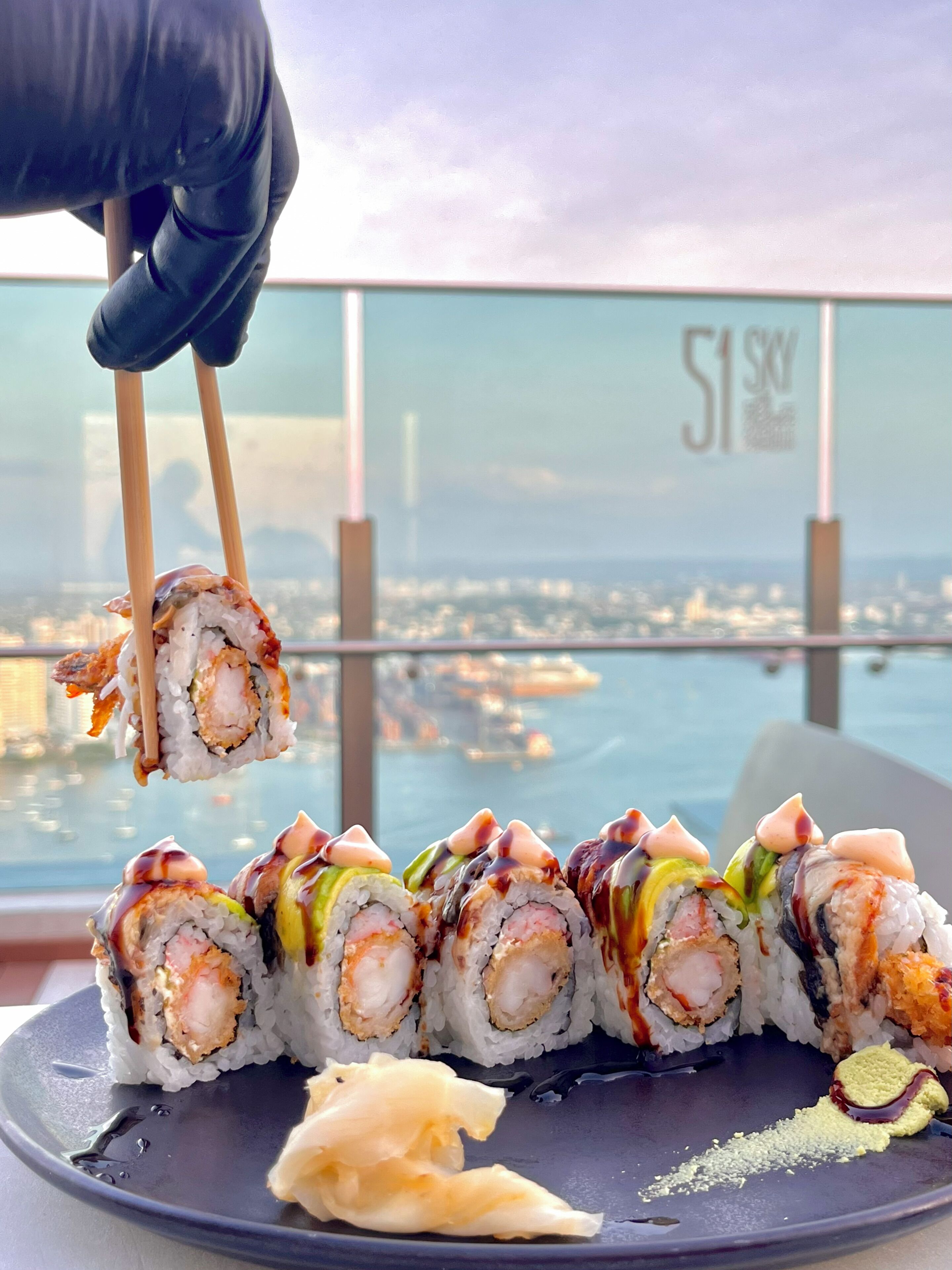 Eine Sushi-Platte, mit Stäbchen, die ein Stück anheben, vor einer Stadtkulisse.