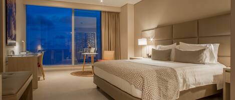 A modern hotel room with a large bed, a desk, and a view of the city at night.