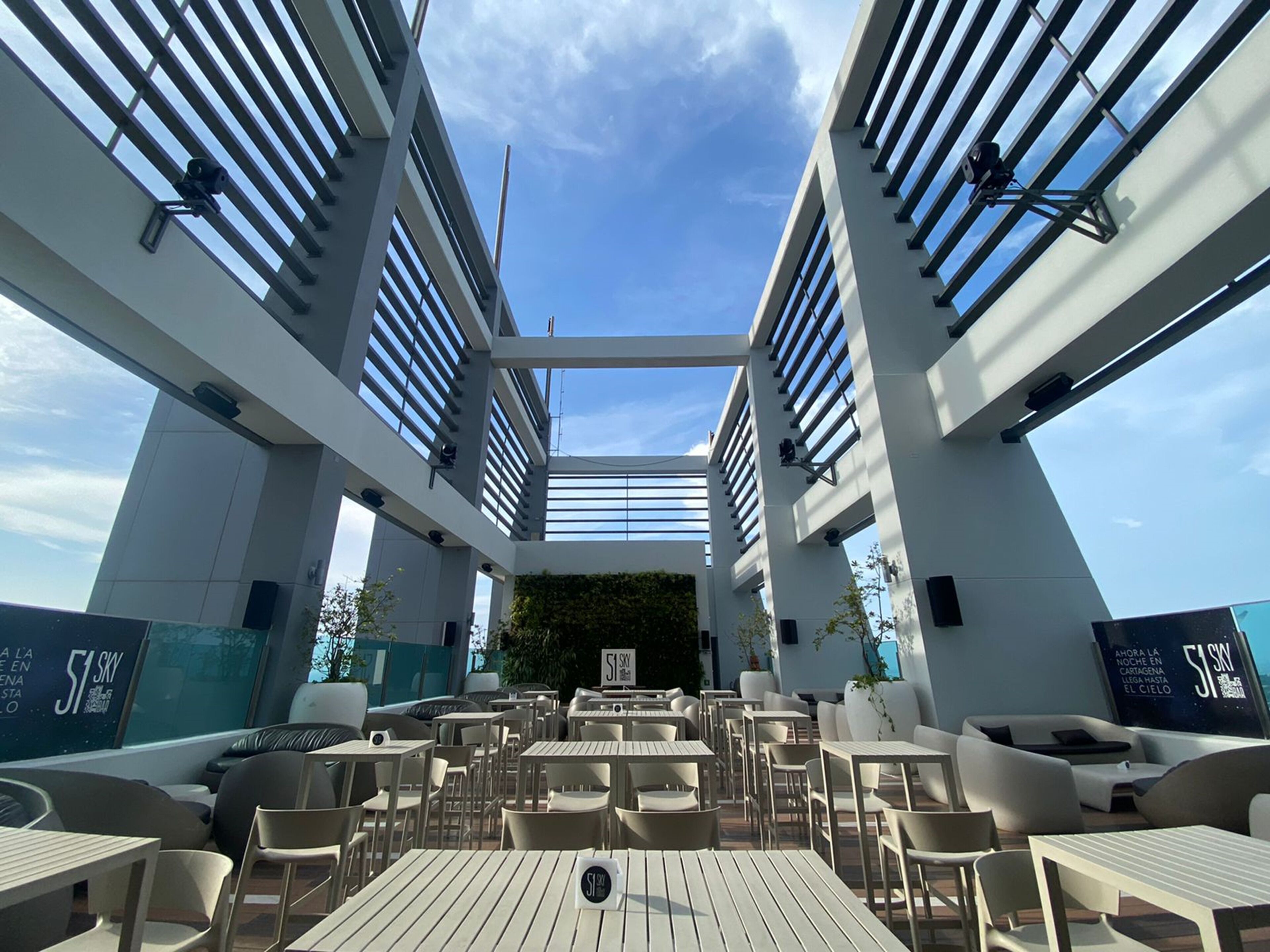 An outdoor seating area with tables and chairs, large windows, and a clear sky.