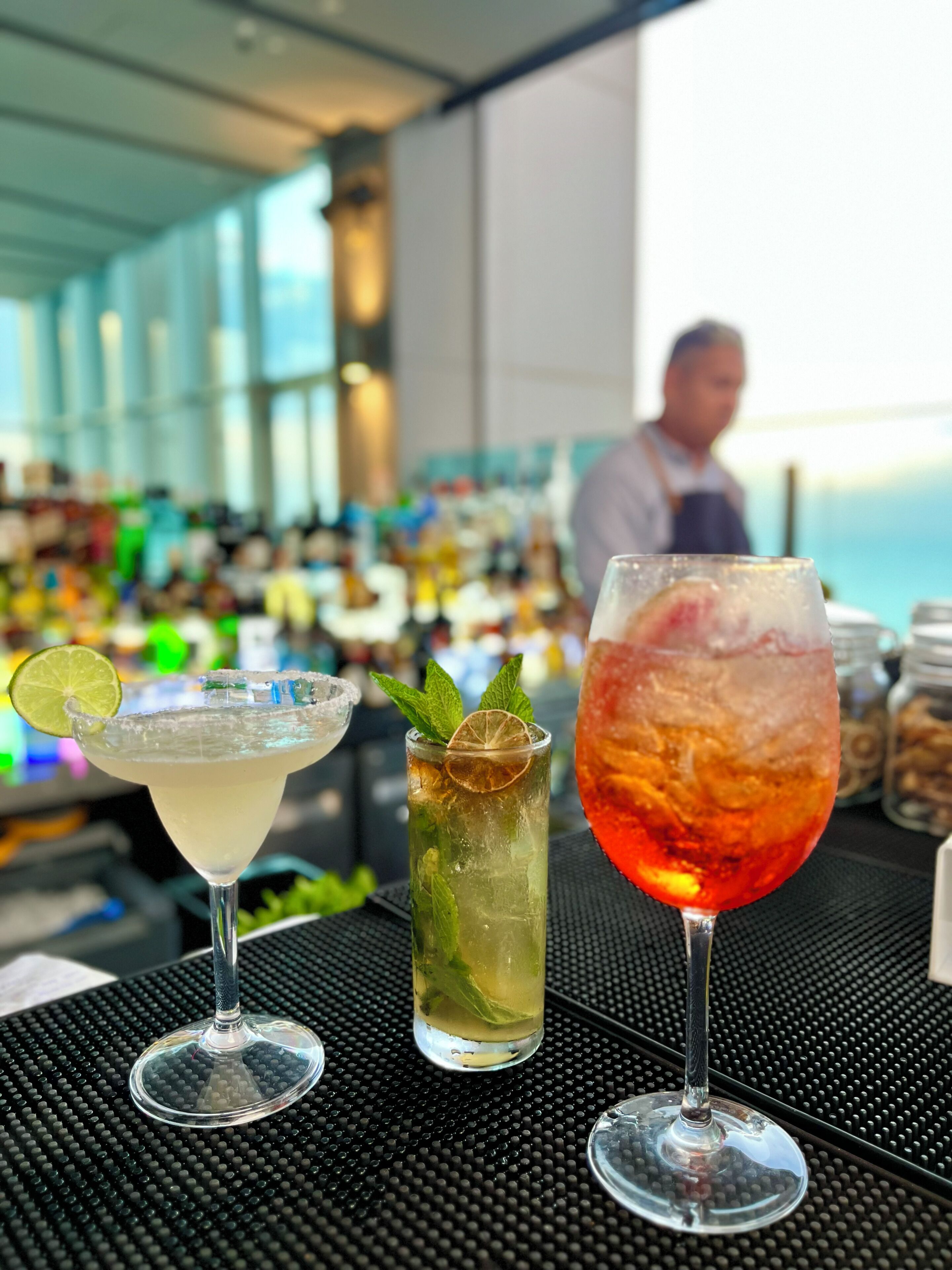 Three cocktails on a bar counter with a bartender in the background.