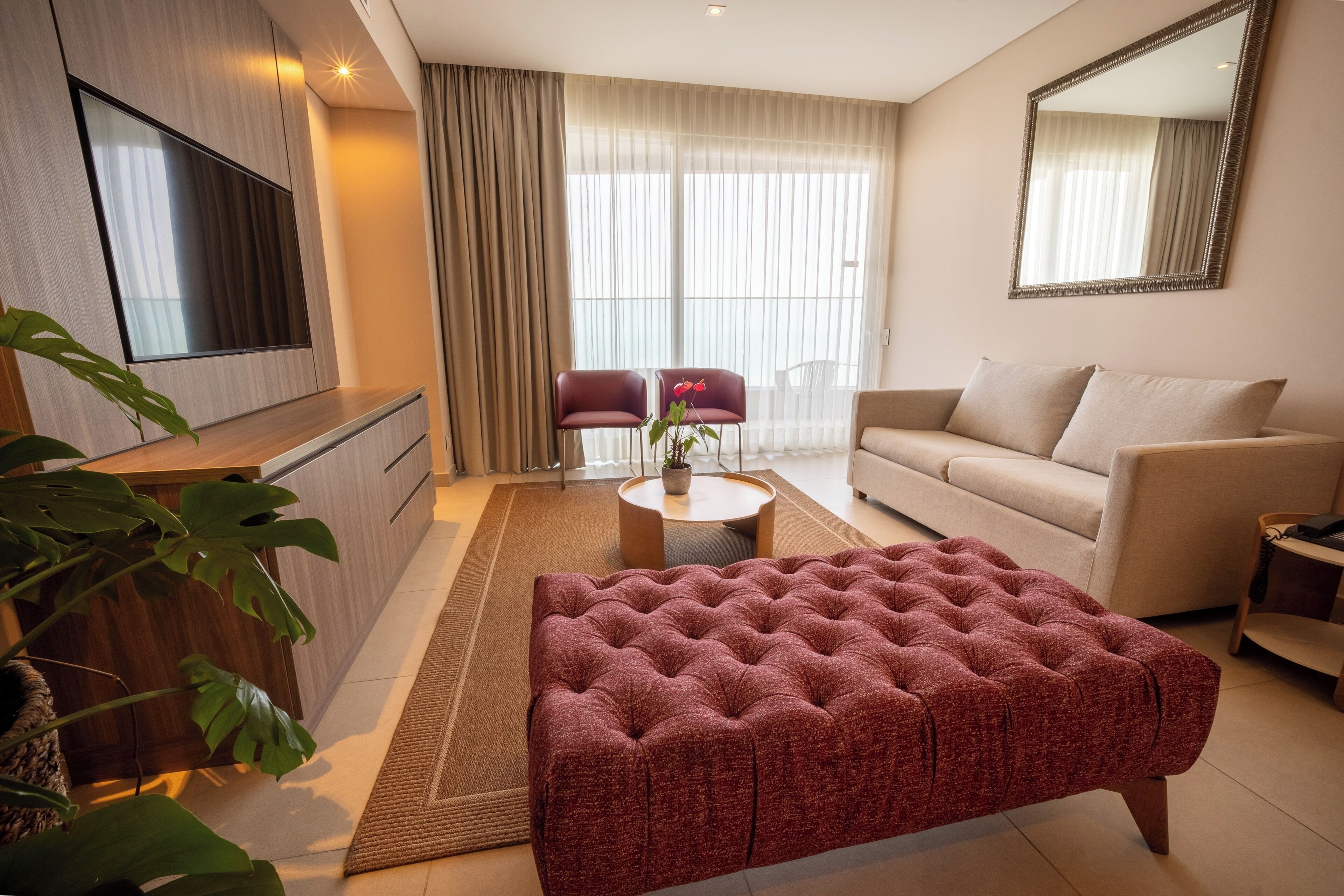 A modern living room with a flat-screen TV, a sofa, a red tufted ottoman, and a large window with curtains.