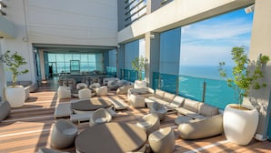 A modern outdoor lounge area with multiple seating options, large windows offering ocean views, and potted plants.