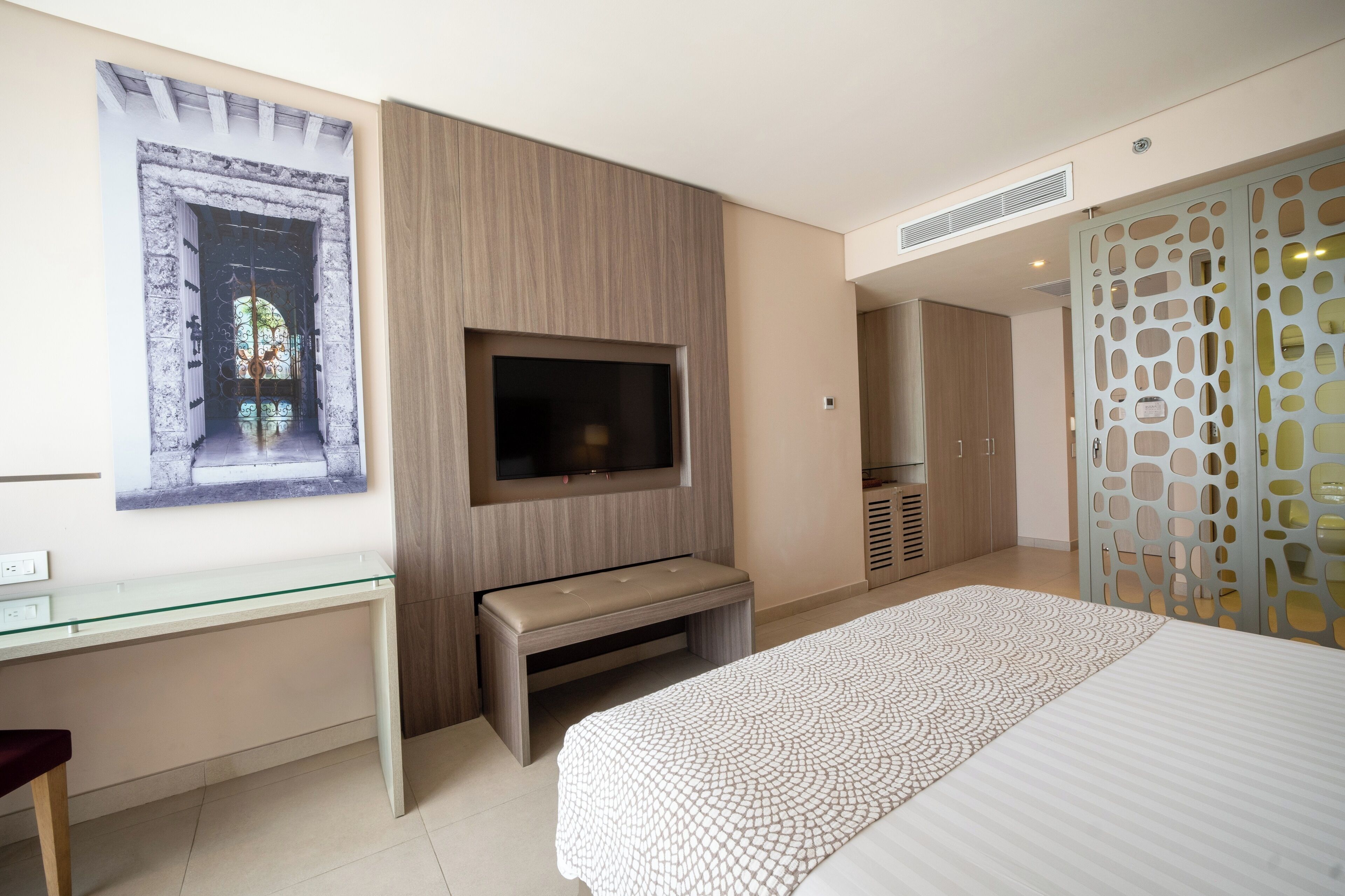 A bedroom with a bed, a TV mounted on the wall, a desk with a bench, and a decorative screen.