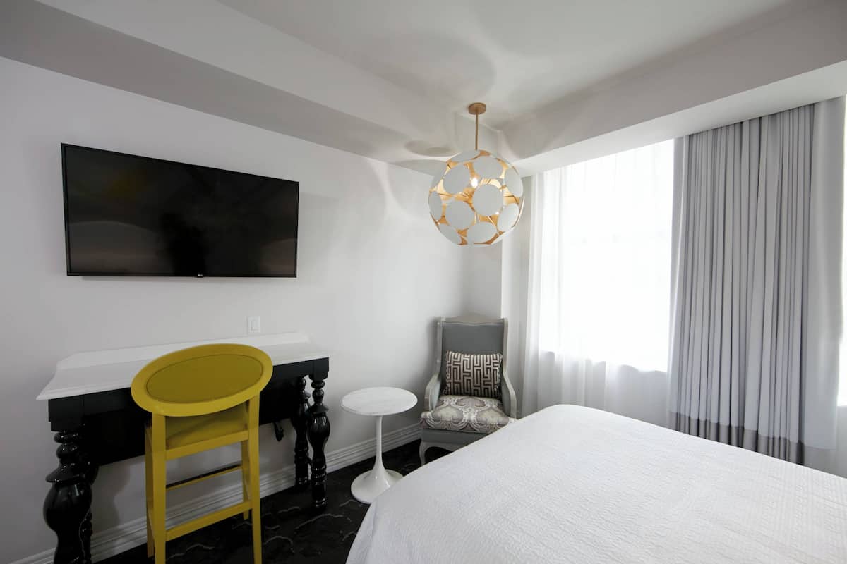 Ein Hotelzimmer mit einem Bett, einem an der Wand befestigten Fernseher, einem Schreibtisch mit Stuhl und einem kleinen runden Tisch.